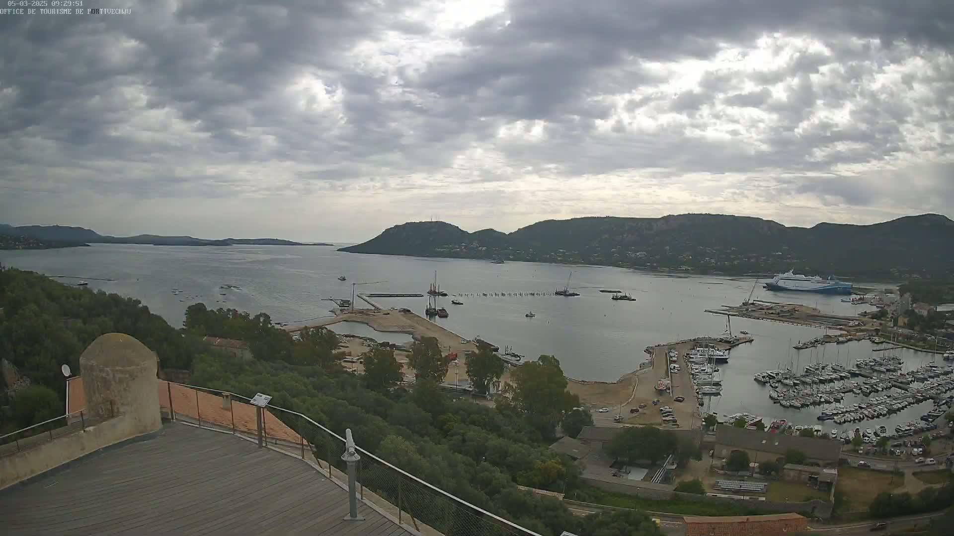 A coastal harbor scene under a cloudy sky shows a large body of water with many boats and ships, including a ferry, docked at a marina near hills and a stone structure.