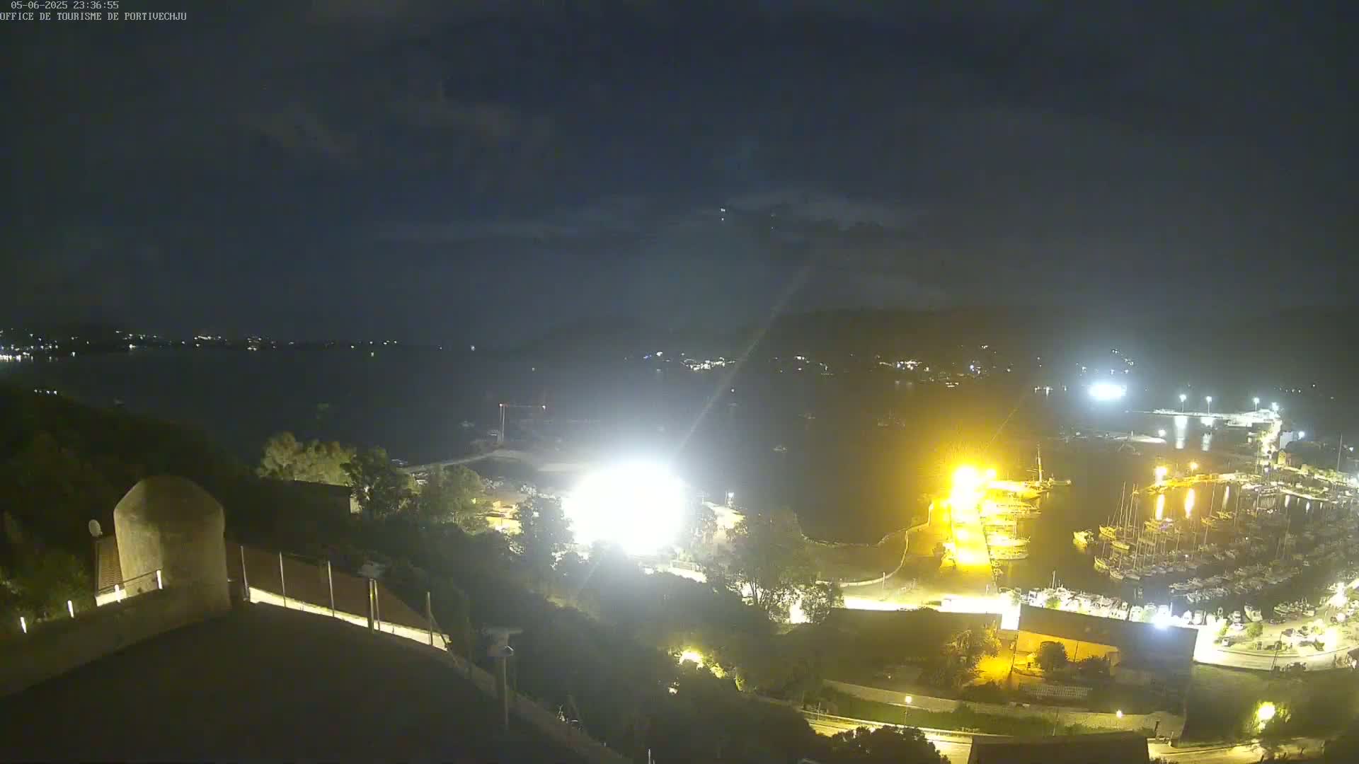 A nighttime view overlooking a harbor filled with boats, brightly lit areas of land, and a dark body of water under a mostly clear sky.