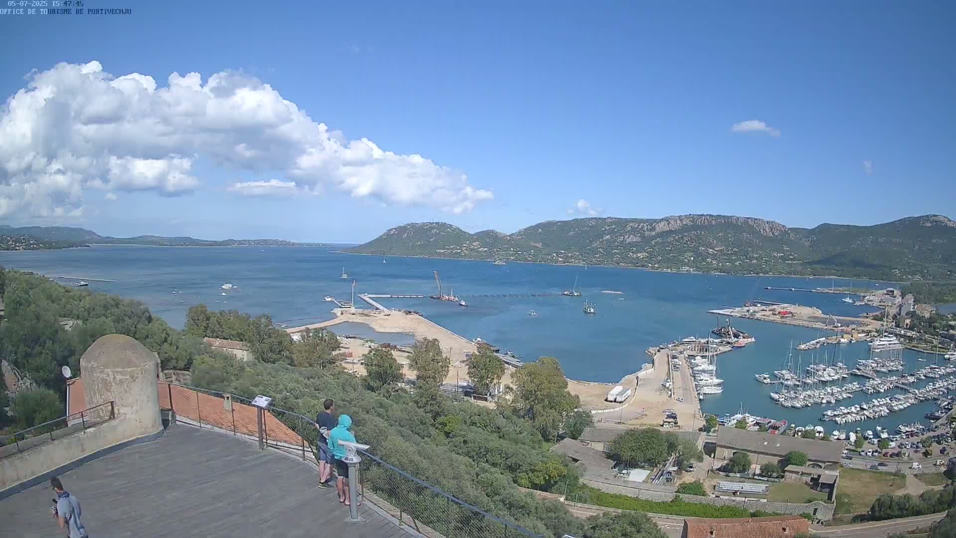 A sunny day shows a coastal harbor filled with boats, viewed from an elevated vantage point, with a few people standing at a railing overlooking the scene.