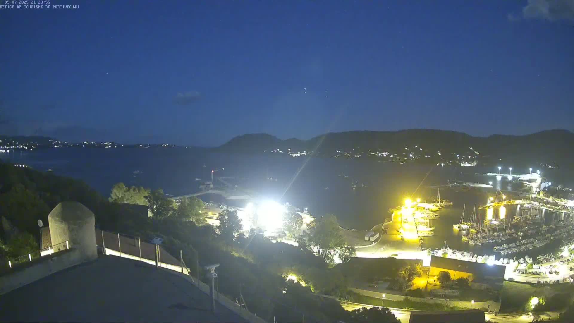 A nighttime view of a harbor filled with boats, under a clear, dark blue sky, with hills visible in the distance.