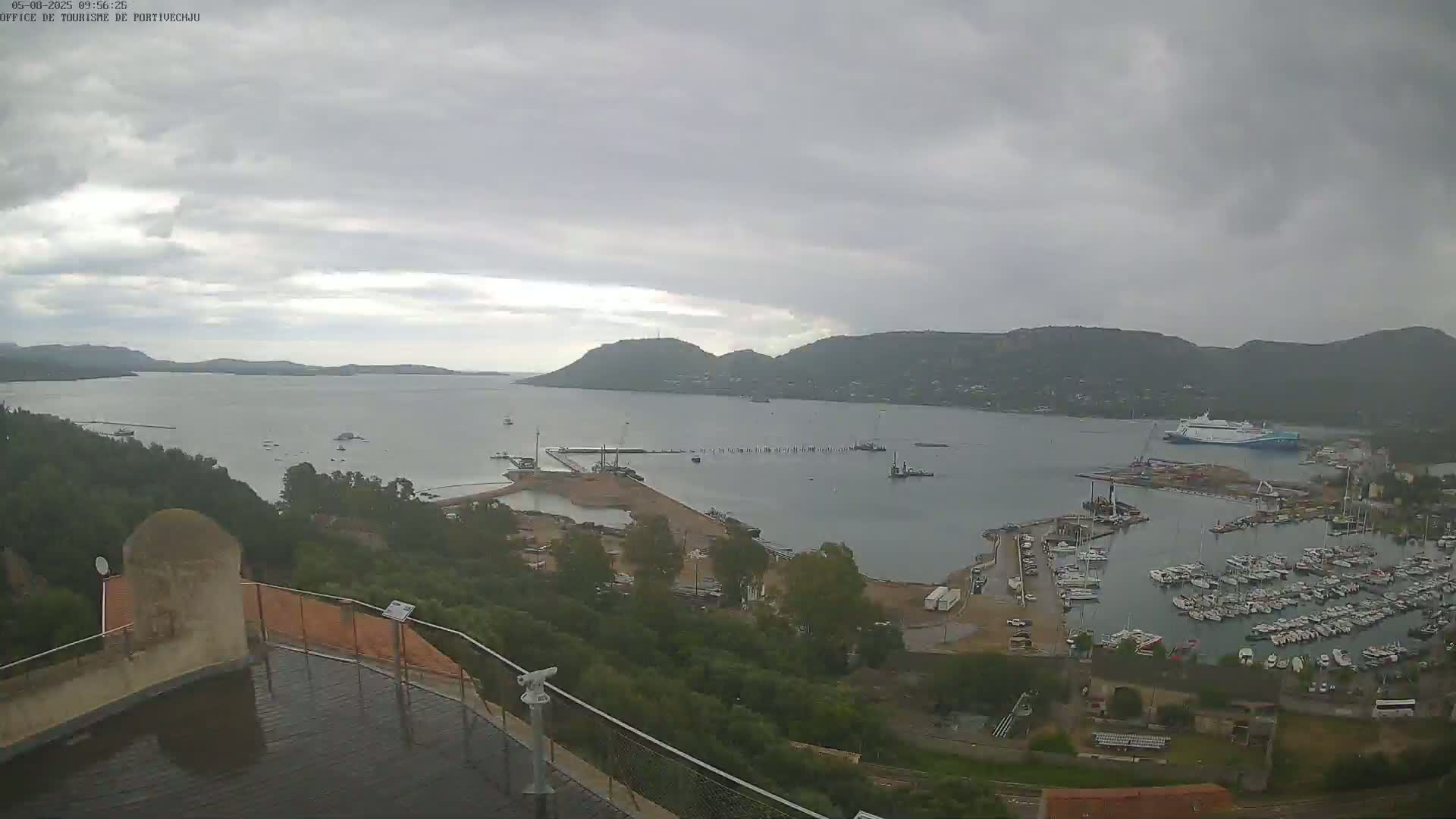 A high-angle, overcast view of a harbor filled with numerous boats and a large ferry, situated between hills and a calm body of water.