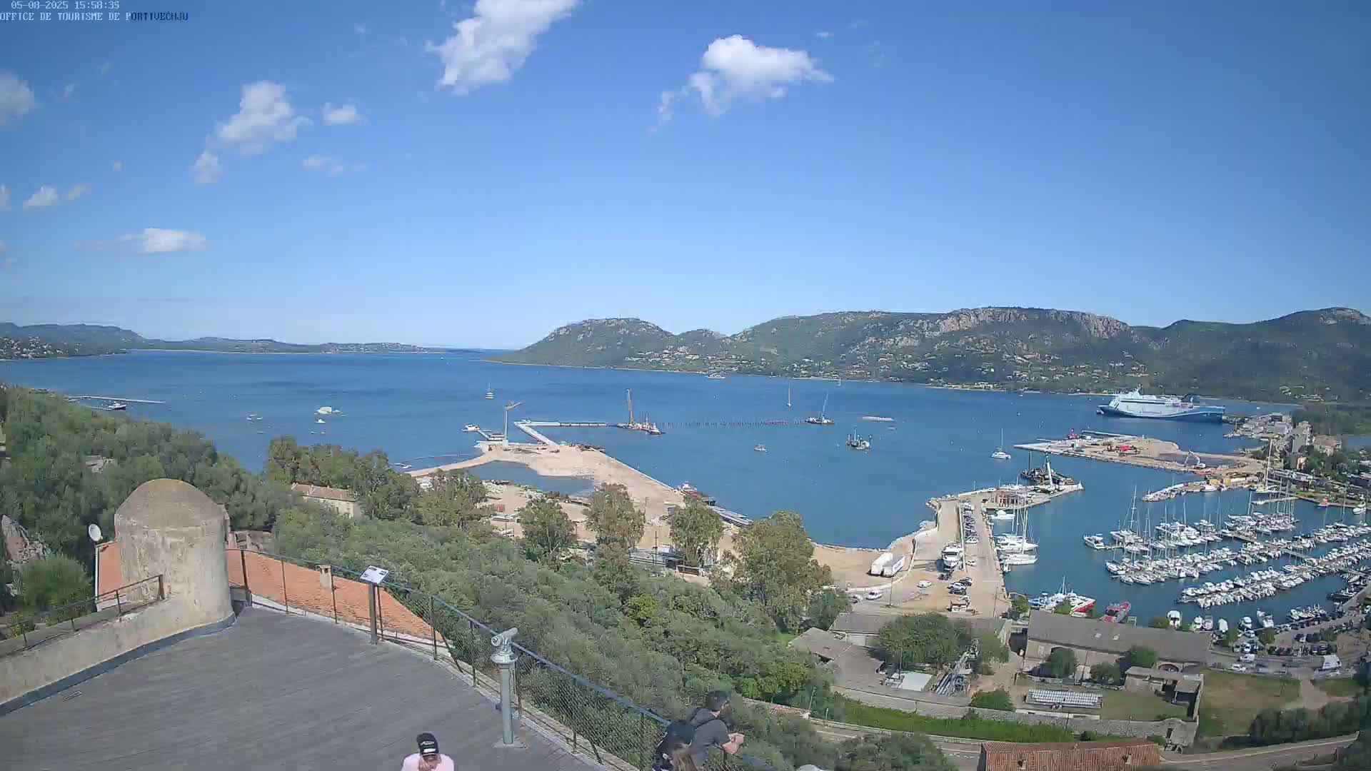 A partly sunny day overlooks a harbor filled with boats, construction, and a large ferry, with hills and a calm bay visible in the distance.