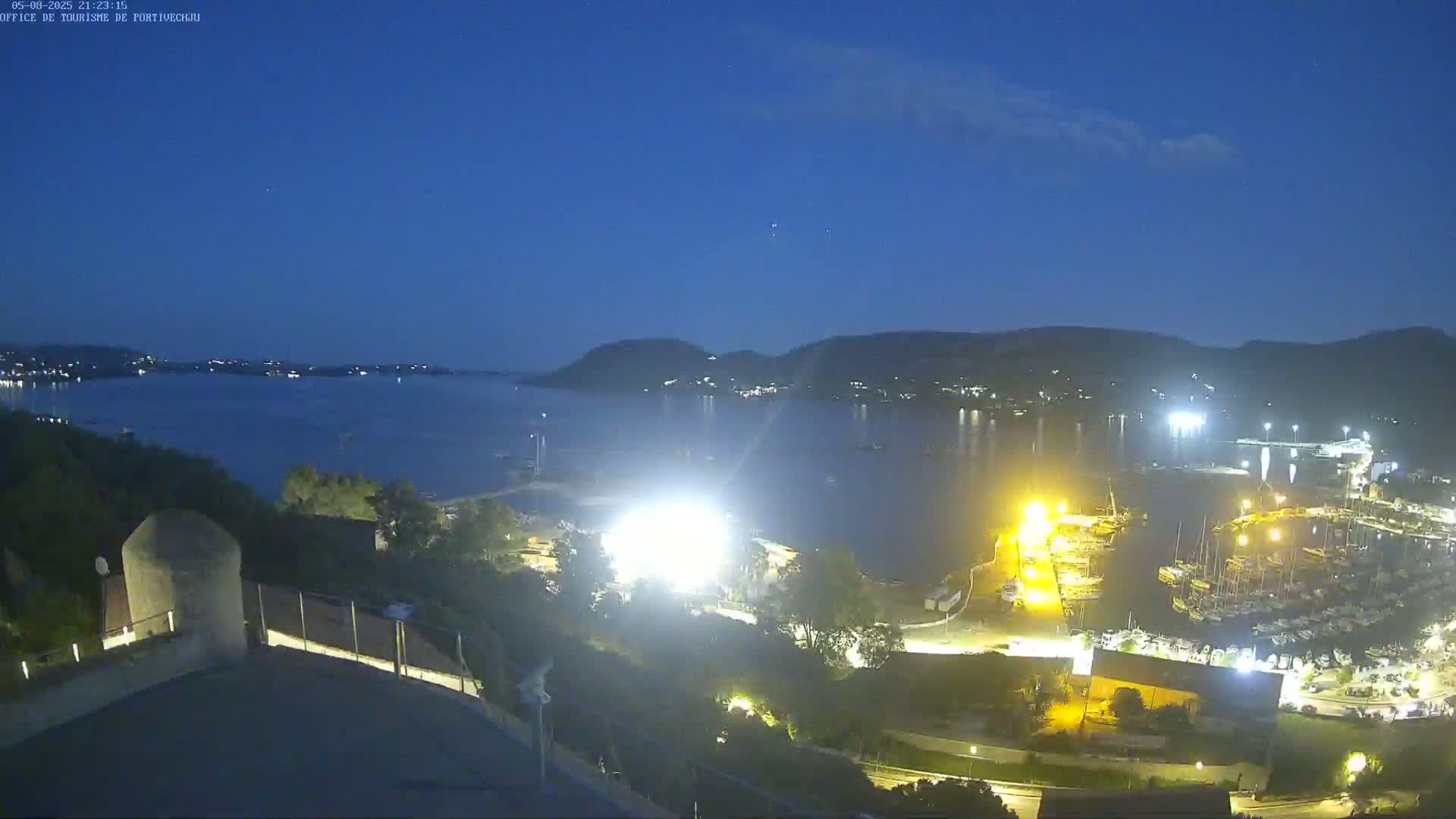 A nighttime view of a harbor filled with boats, illuminated by lights, with hills in the background under a clear, dark blue sky.