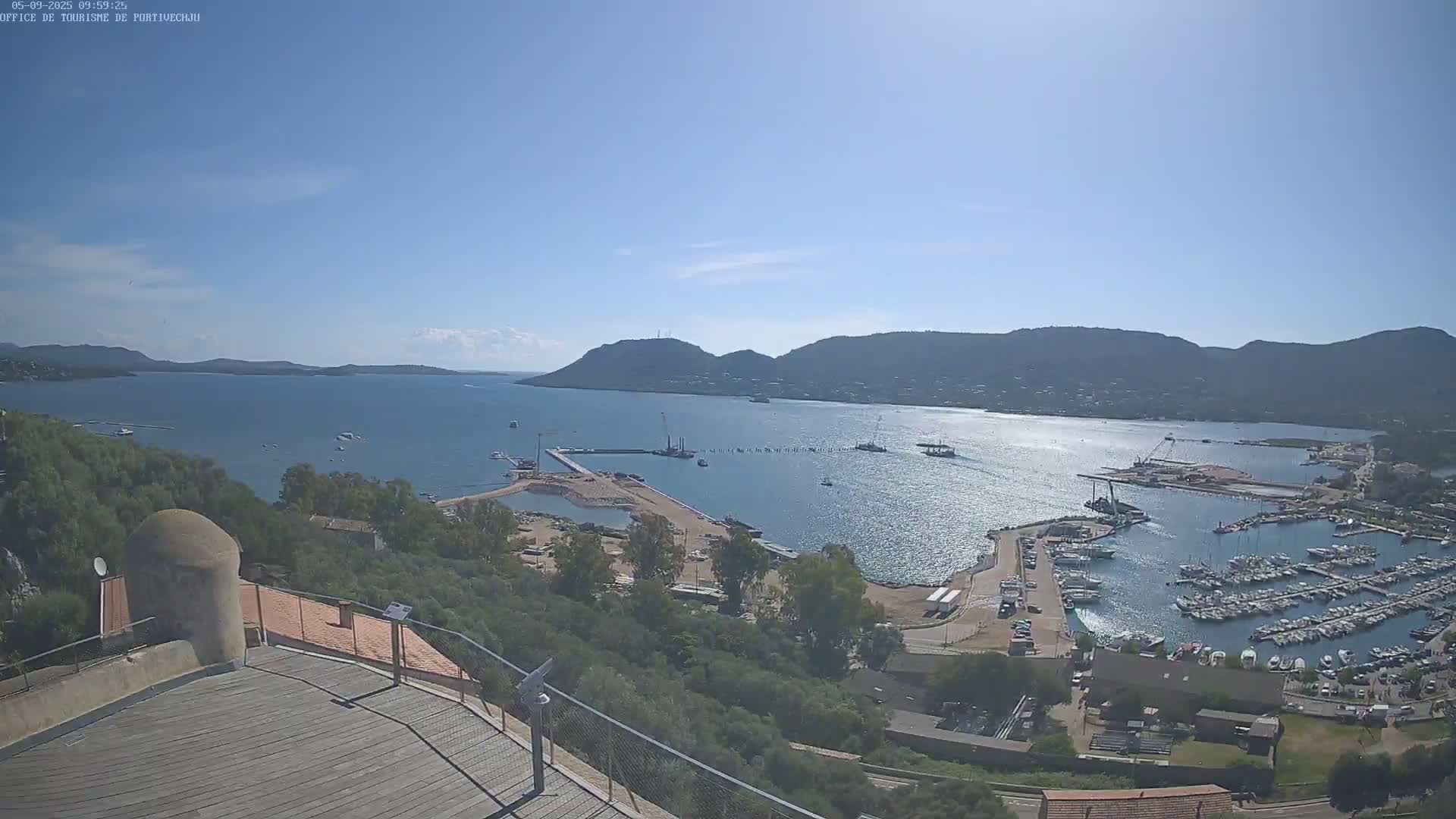 A high-angle view on a sunny day shows a harbor filled with boats and construction activity, set against a backdrop of mountains and calm blue water.