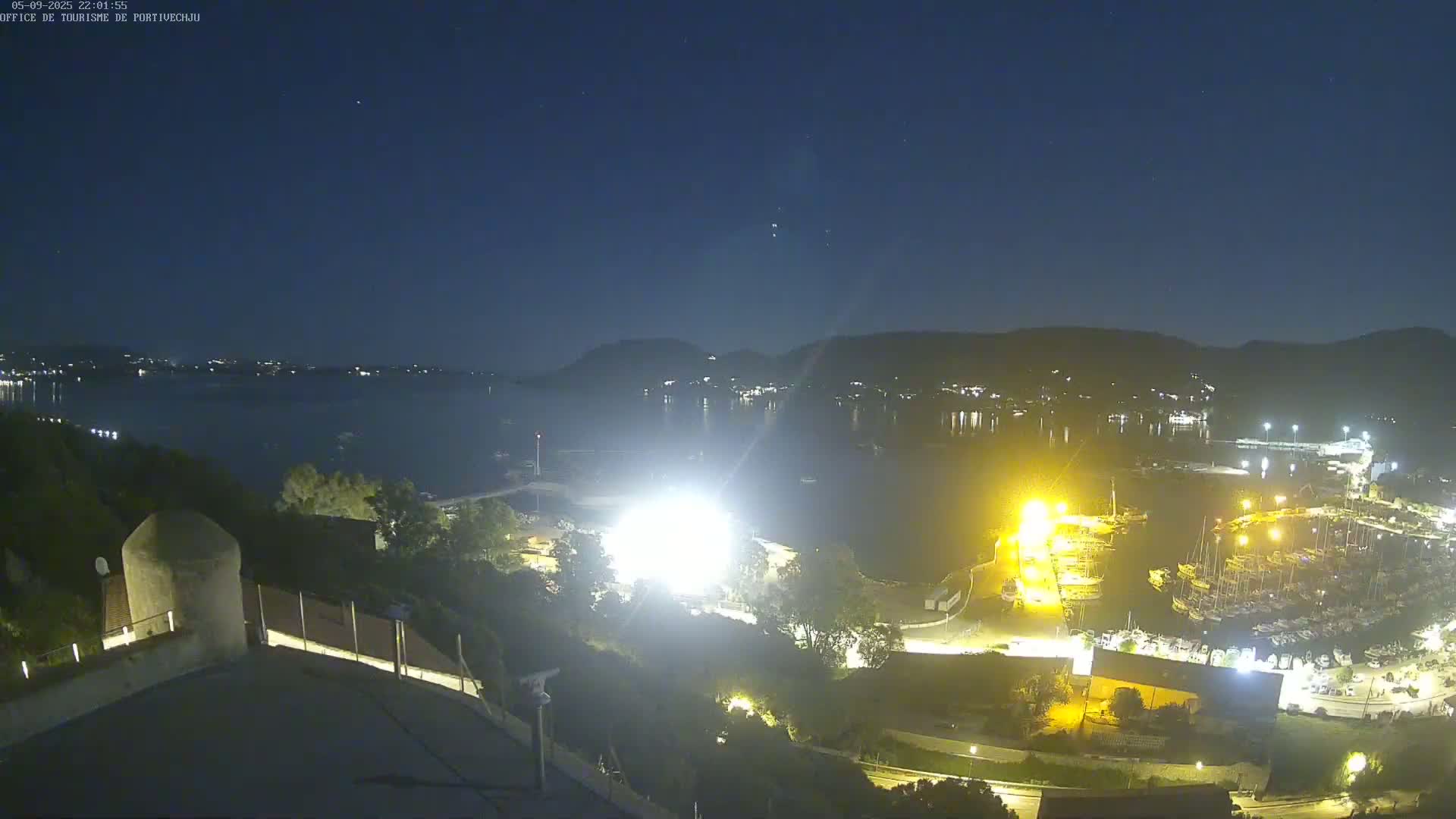 A nighttime, high-angle view of a harbor filled with boats, illuminated by bright lights, with a calm body of water extending to a dark hillside in the background under a clear, starry sky.
