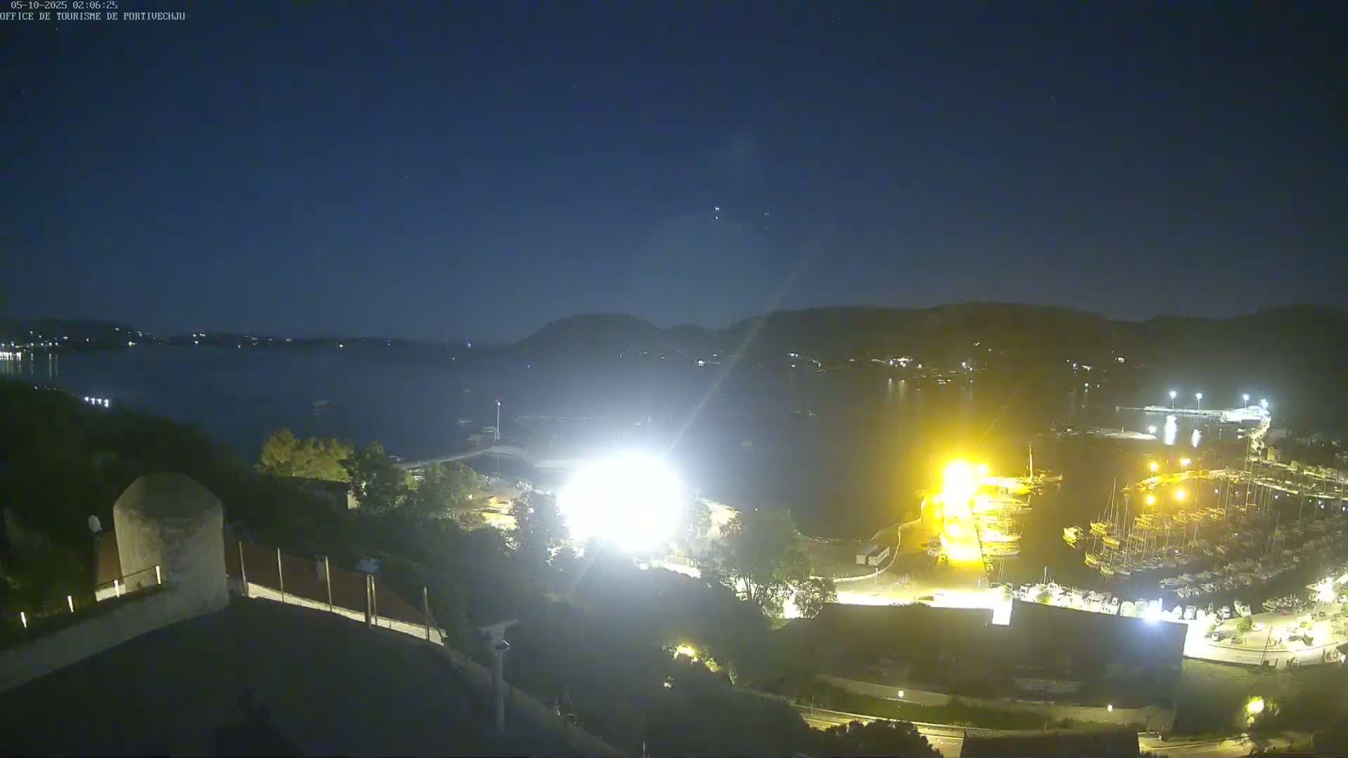 A nighttime view of a marina filled with boats, brightly lit, overlooking a dark body of water under a clear, starlit sky.