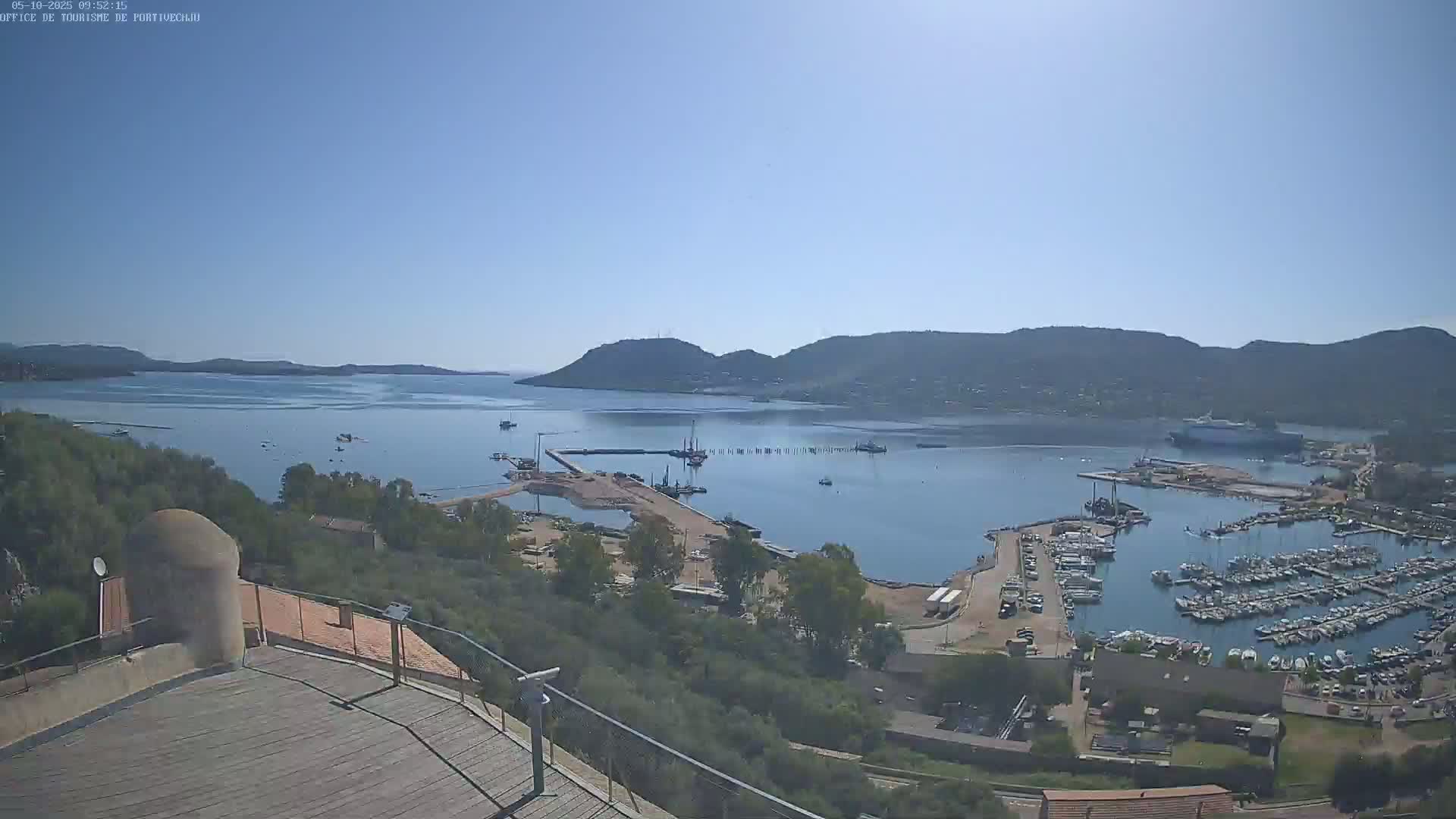 A high-angle view shows a calm, sunny day over a harbor filled with boats, surrounded by green hills and a clear blue sky.