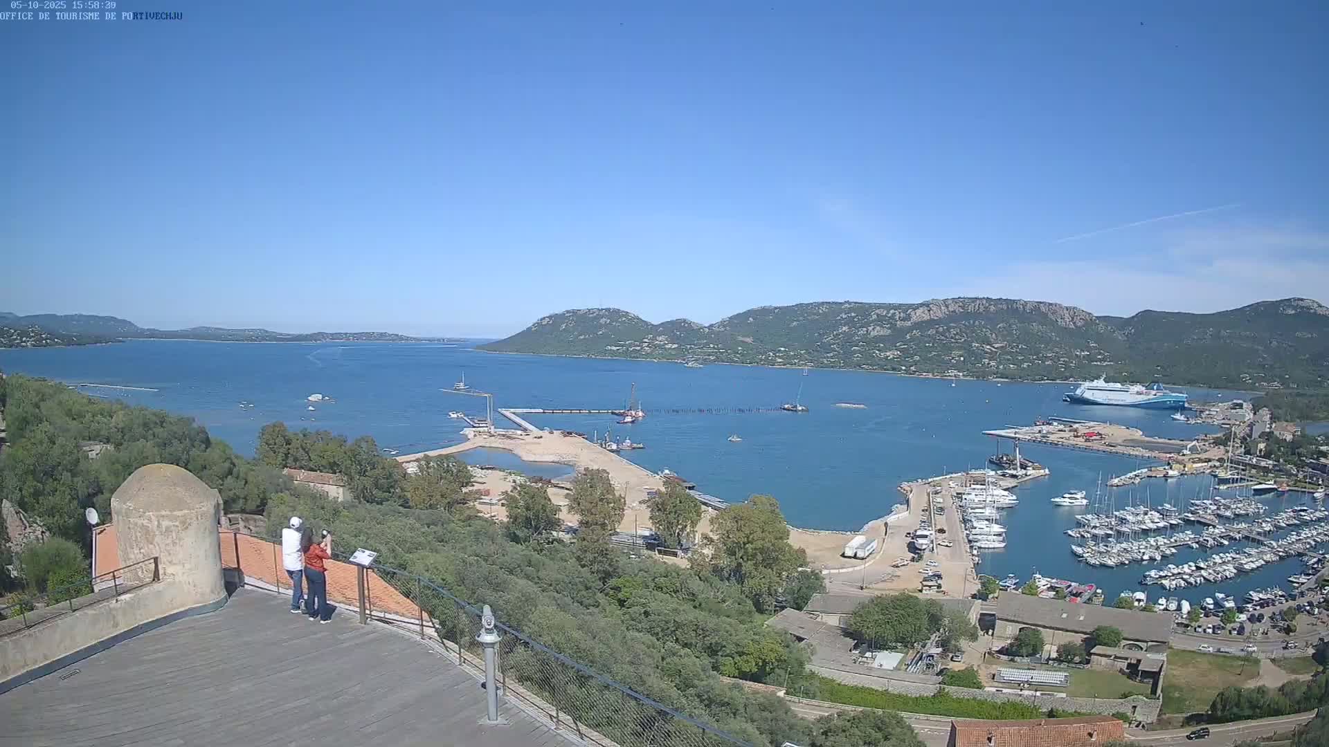 A panoramic view from a high vantage point shows a harbor filled with boats under a clear blue sky, with hills in the distance and two people observing the scene from a stone structure.