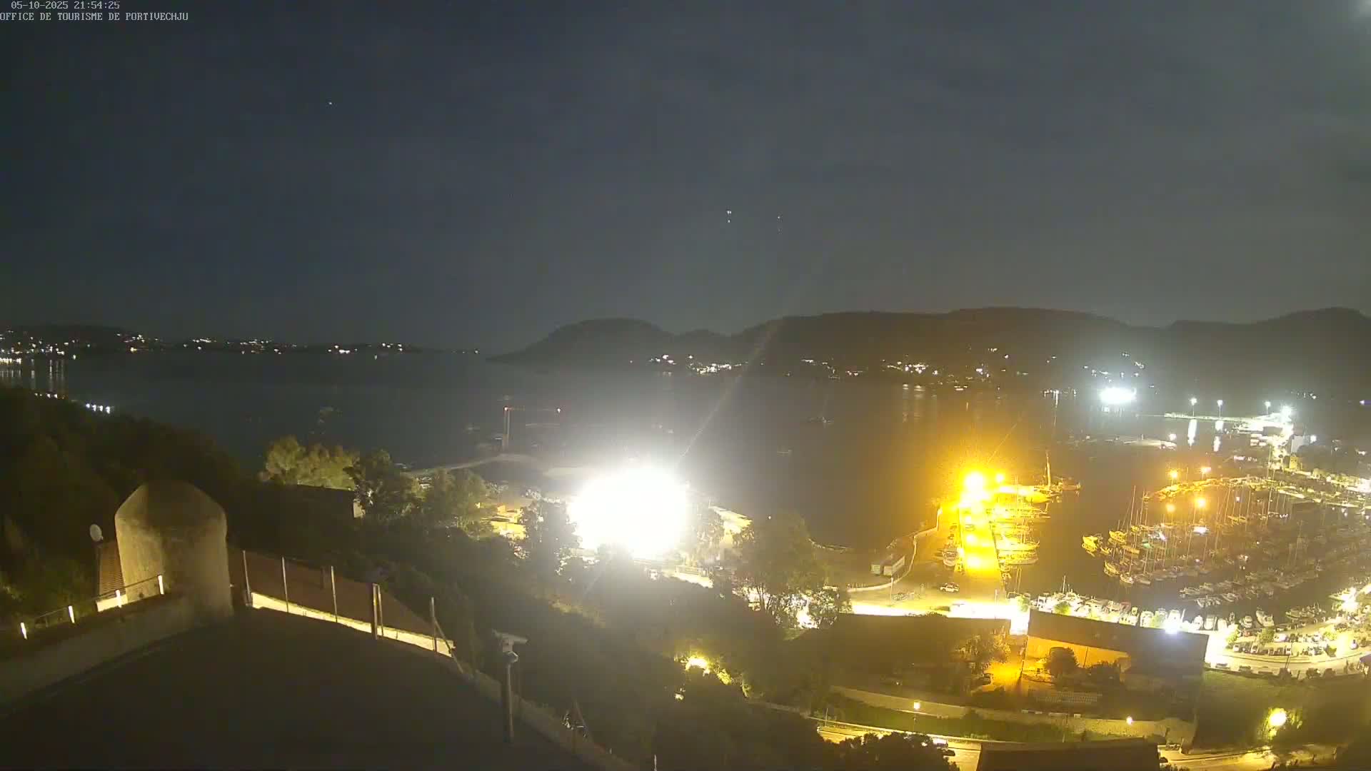A nighttime, high-angle view shows a body of water with a marina filled with boats, hills in the background, and a brightly lit area on the shore.