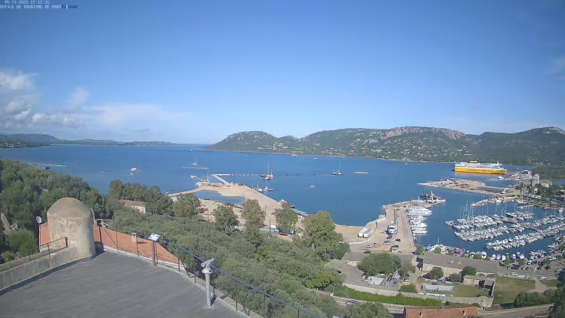 A high-angle, long shot on a clear day shows a harbor filled with boats, a large yellow ferry, and construction activity near a calm body of water surrounded by hills and vegetation.