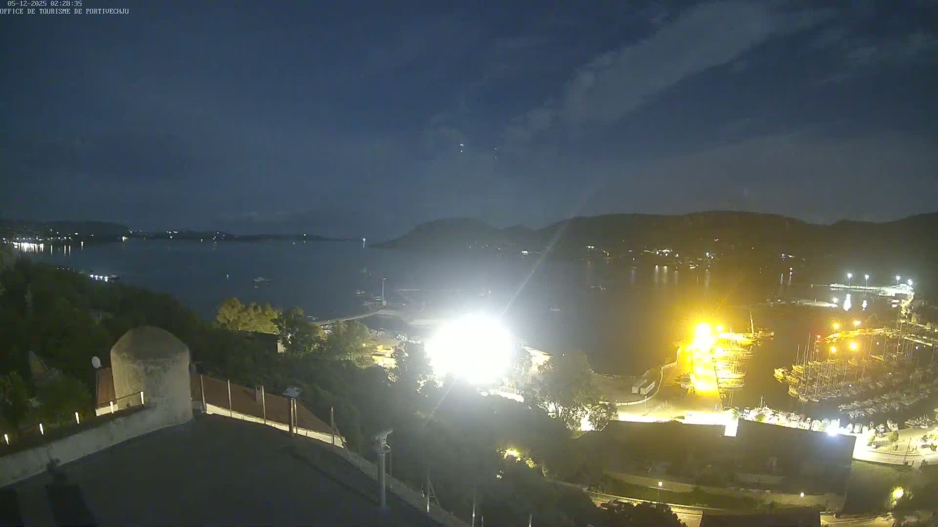 A nighttime view of a calm bay with a marina filled with boats, lit by artificial lights under a partly cloudy sky.