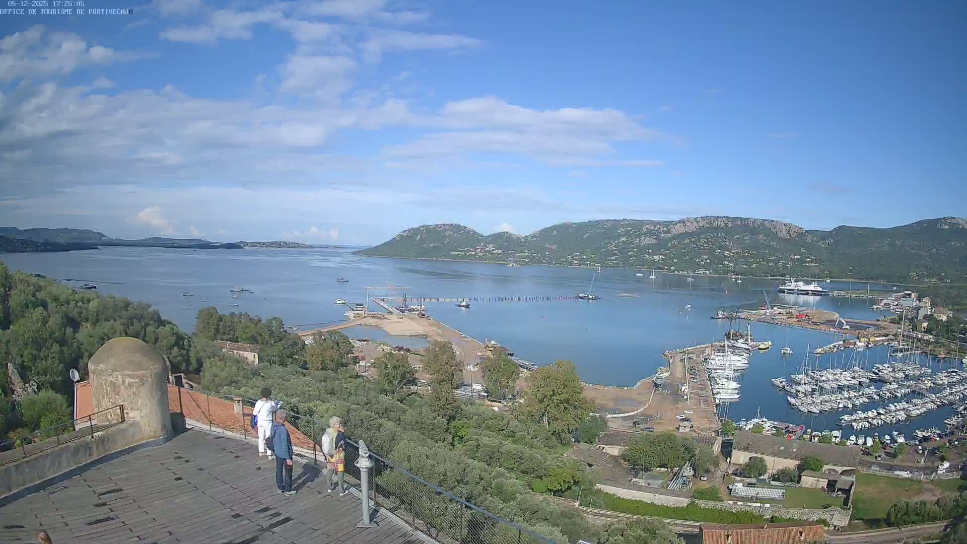 A picturesque harbor filled with sailboats and yachts is seen from an elevated vantage point on a partly cloudy day, with a calm bay and distant hills in the background.