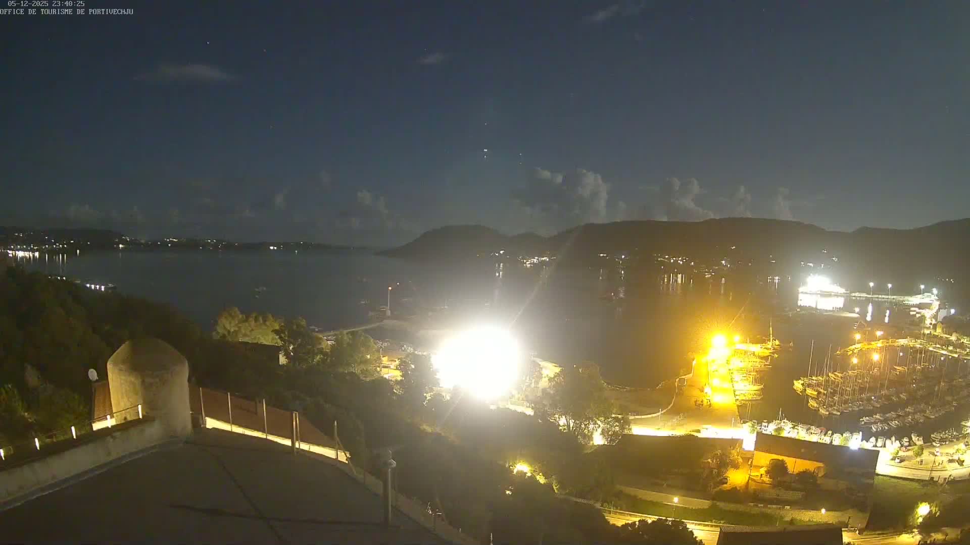 A nighttime, high-angle view of a harbor filled with boats, illuminated by bright lights, with a calm body of water extending to dark hills under a mostly clear sky.