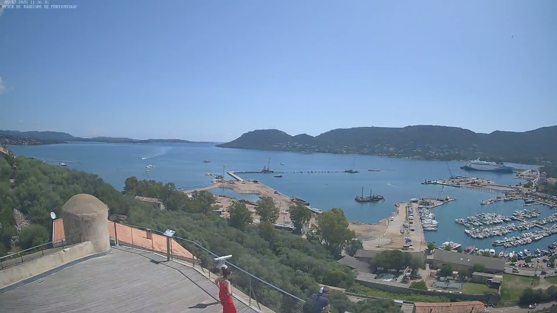 A sunny day overlooking a harbor filled with boats, a partially-constructed dock, and a calm bay surrounded by hills.
