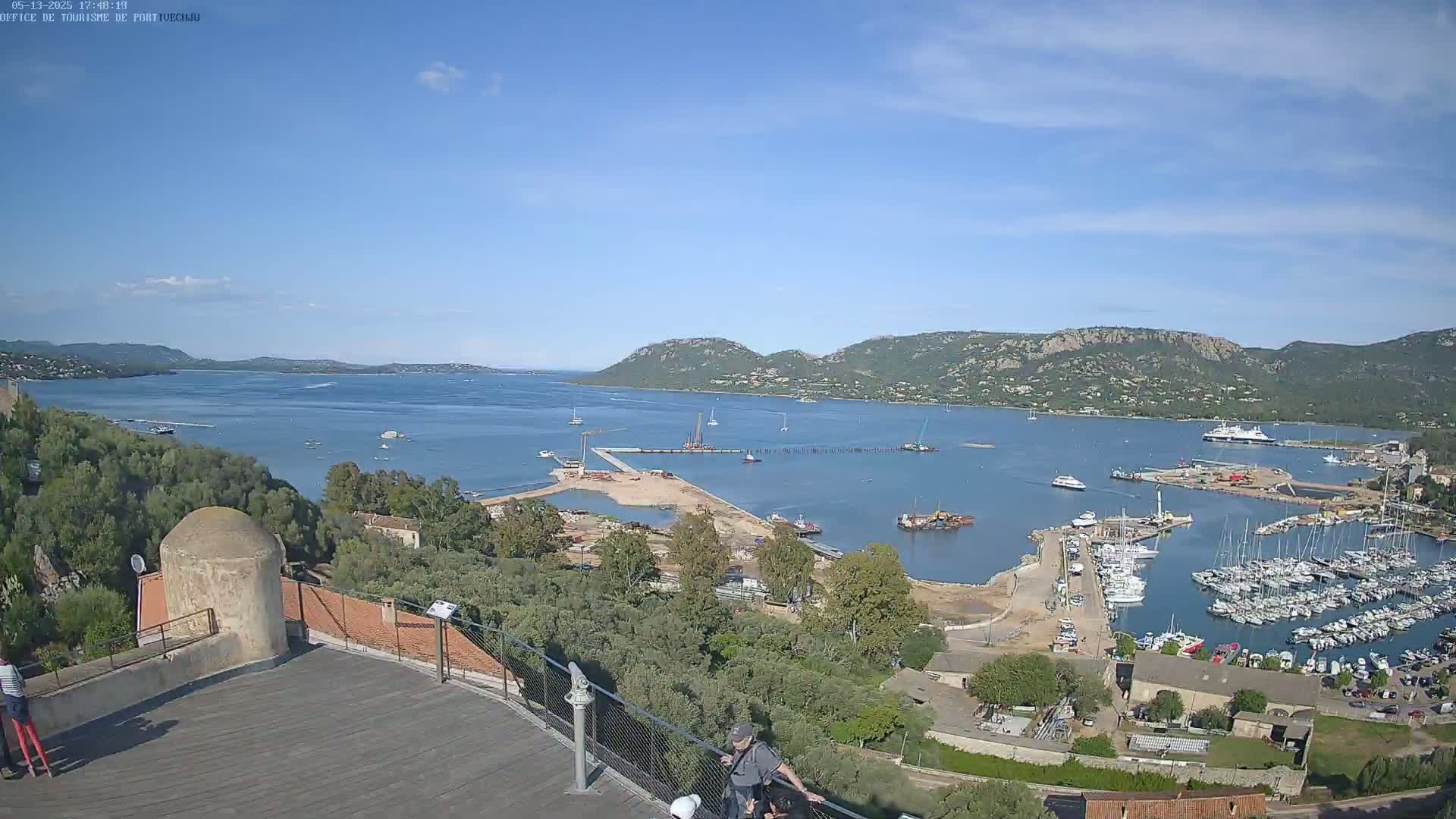 An expansive view of a harbor filled with sailboats and yachts, under a mostly clear blue sky, with hills and a calm bay visible in the distance.