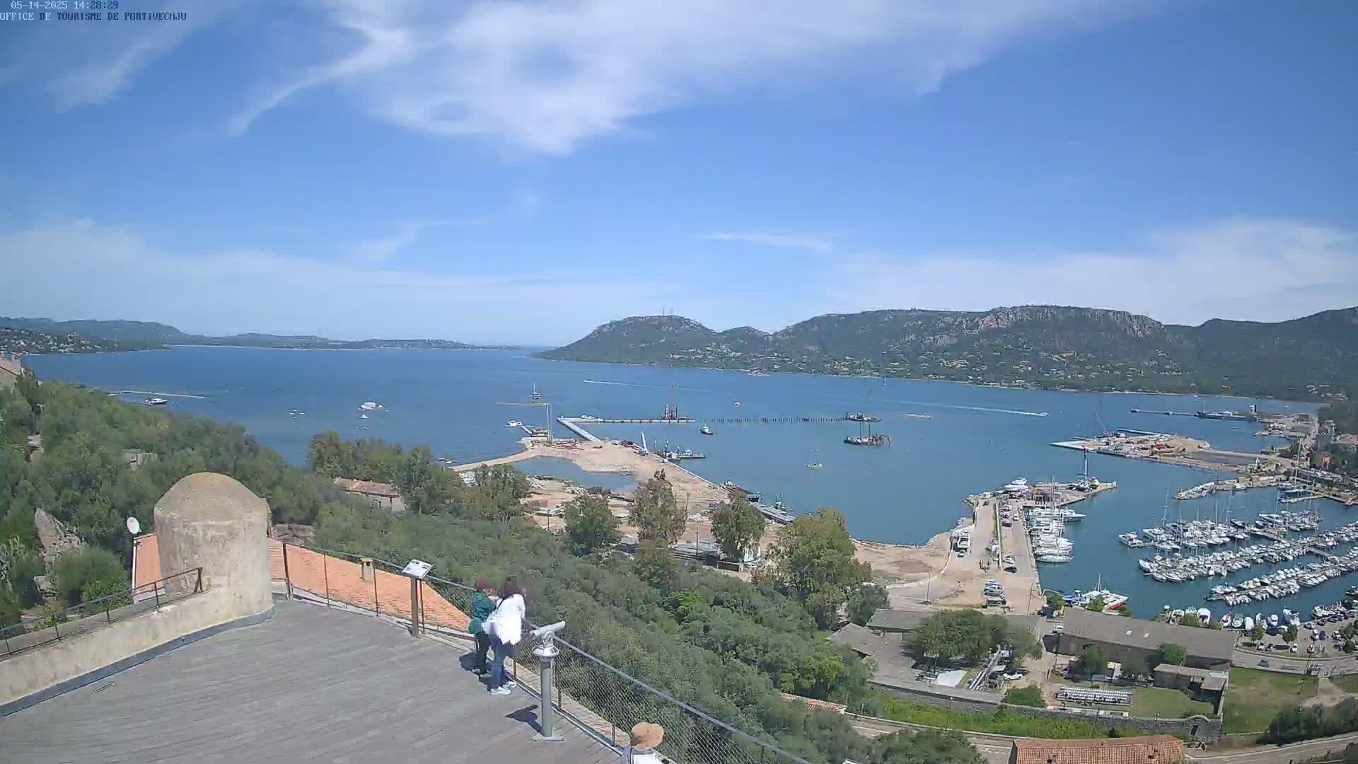 An elevated view shows a partly sunny day overlooking a harbor filled with boats, construction activity, and a calm bay extending to distant mountains under a mostly clear sky.