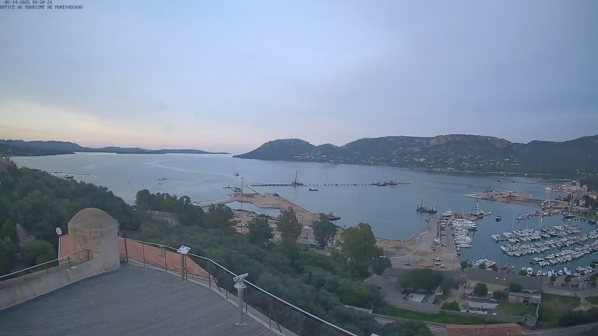 A high-angle, long shot shows a calm bay with many boats in a marina, under a clear, pale-blue sky, with hills and greenery in the background.