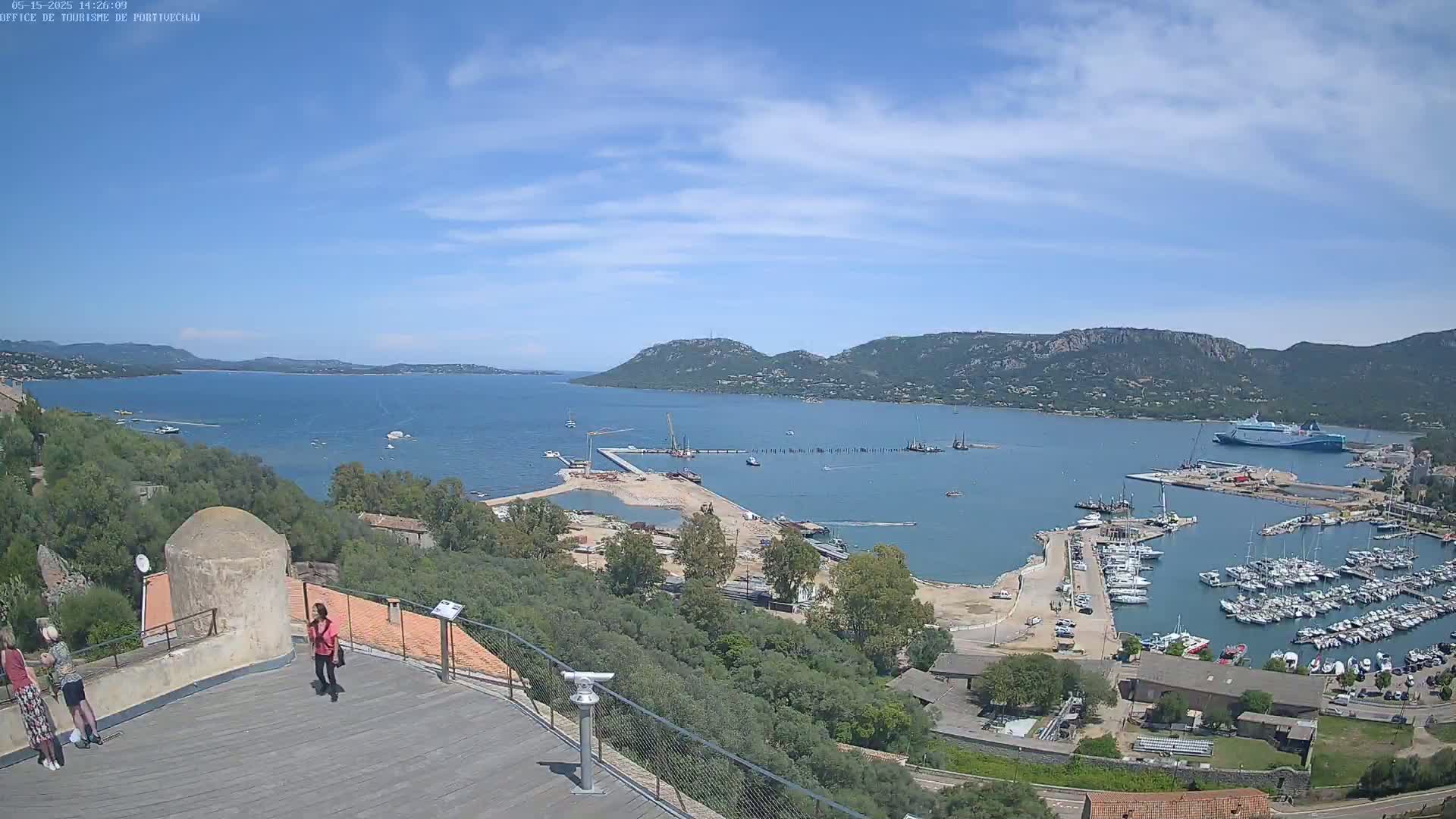 A sunny day overlooking a harbor filled with boats, construction, and a large ferry, with hills and calm blue water in the background, viewed from a high vantage point with a few people present.