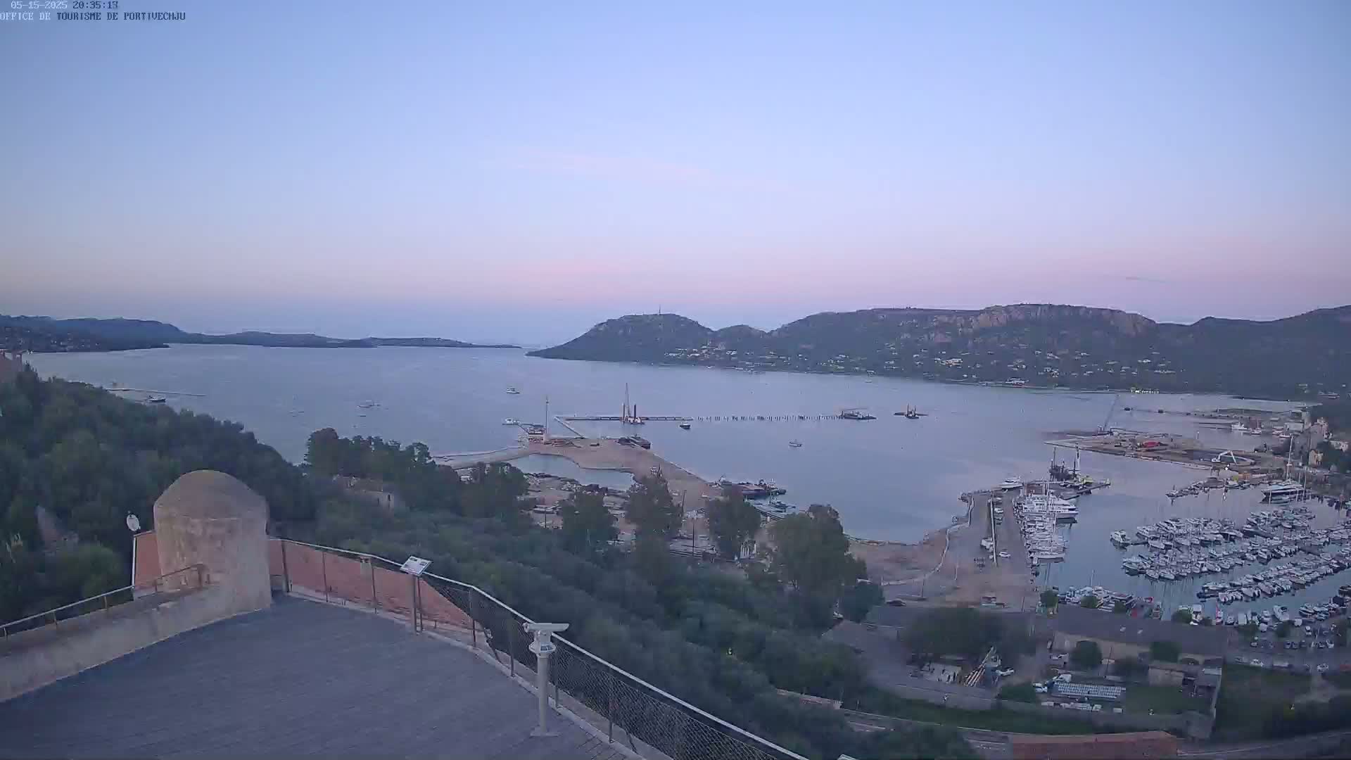 A high-angle, long shot shows a calm bay with many boats docked in a marina, hills in the background under a twilight sky.