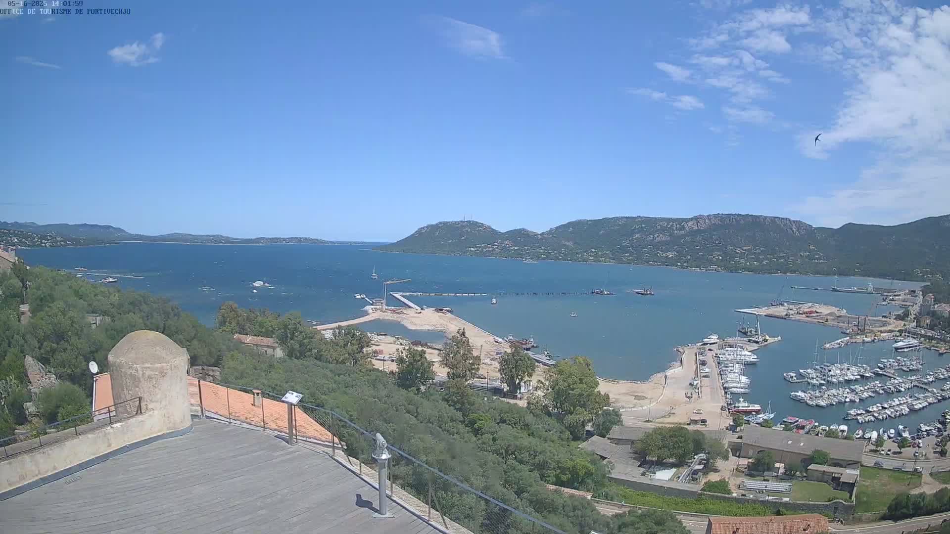 An elevated view shows a calm, sunny day overlooking a harbor filled with boats, a partially constructed area, and a bay surrounded by hills and greenery.