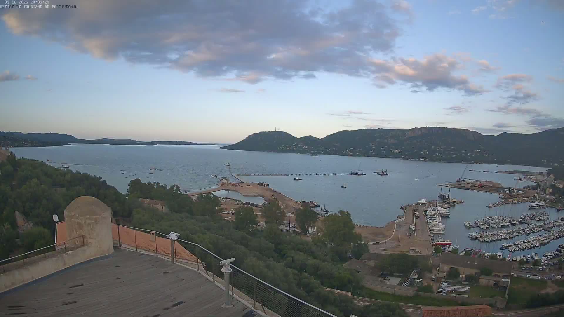 An expansive view of a calm bay filled with numerous boats in a marina, overlooked by green hills under a partly cloudy sky.