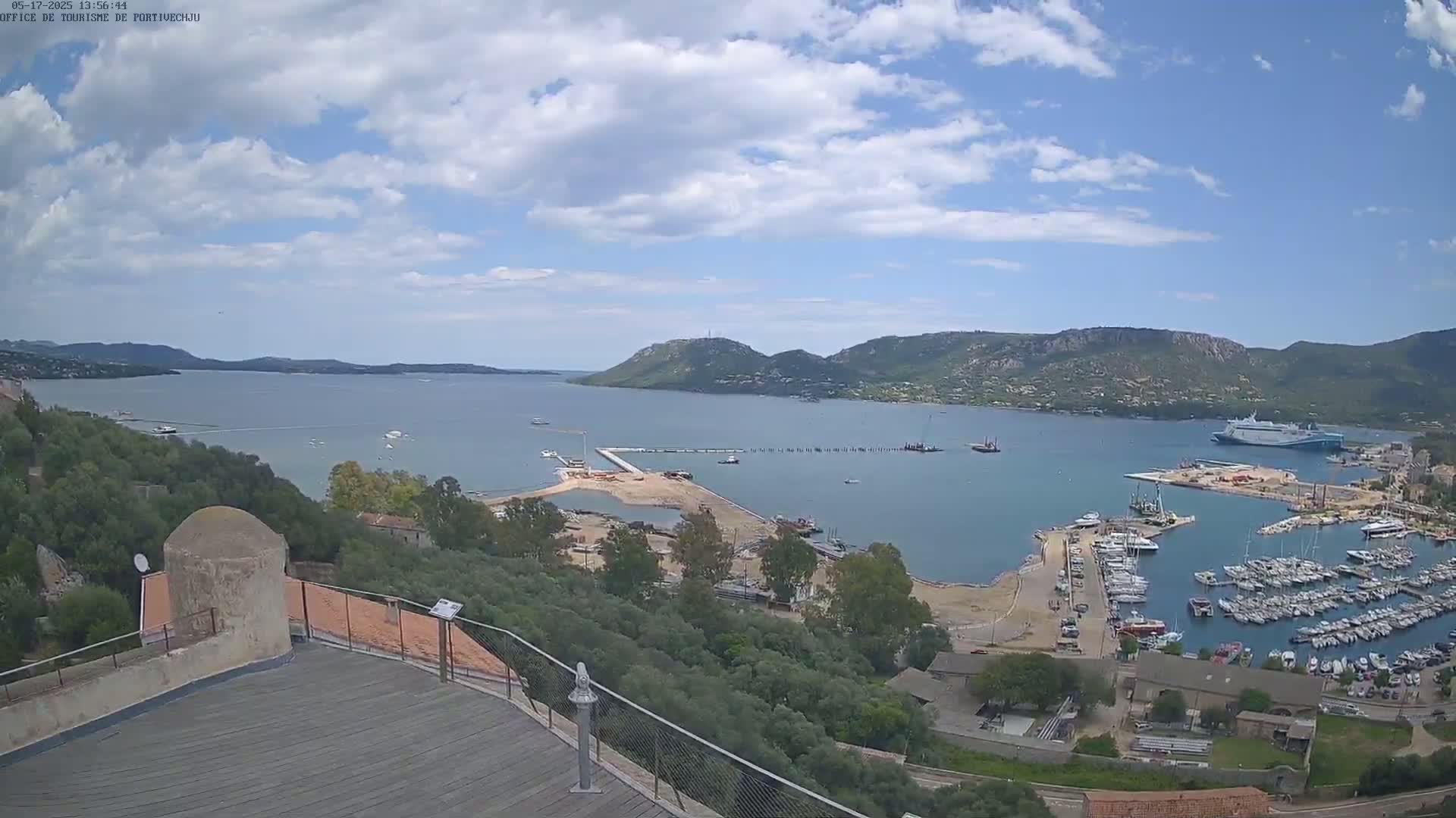 A partly cloudy day overlooks a harbor filled with numerous boats and a large ferry, with ongoing construction visible near the shoreline and hills in the distance.