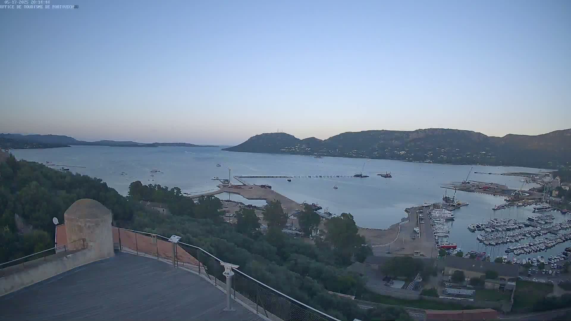 An elevated view shows a calm bay with numerous boats docked at a marina, surrounded by hills under a clear, twilight sky.