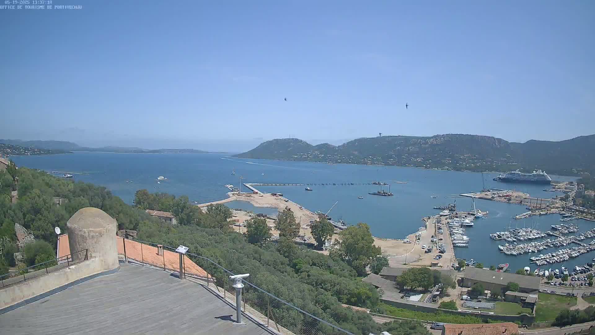 An elevated view of a harbor filled with numerous boats and a large cruise ship, under clear blue skies, shows a construction site near the water and hills in the background.