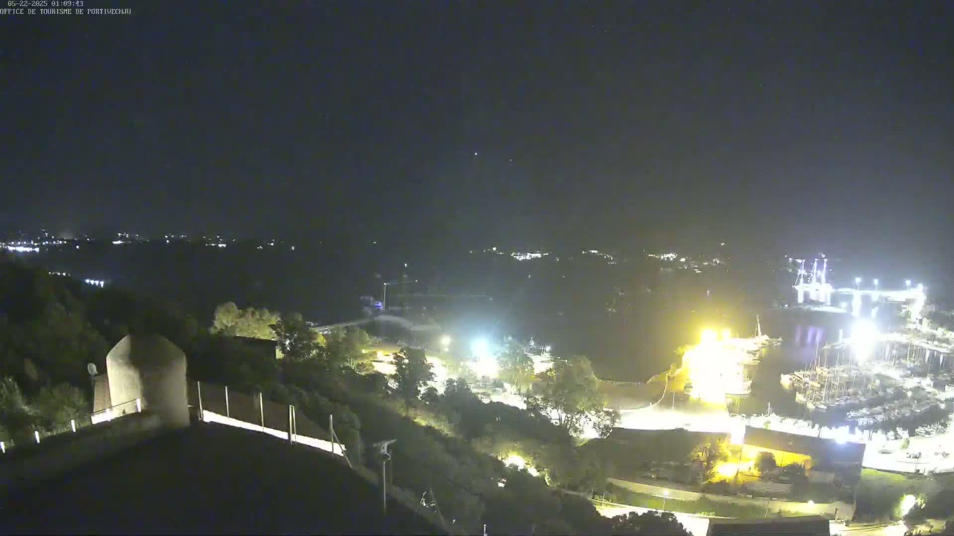 A nighttime, high-angle view shows a marina with many boats, a town in the distance, and a hilltop structure illuminated by lights.
