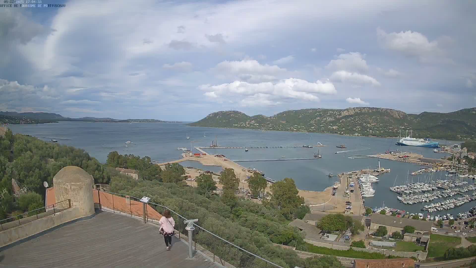 A woman walks along a wooden deck overlooking a harbor filled with boats, under a partly cloudy sky, with hills and a large body of water in the distance.