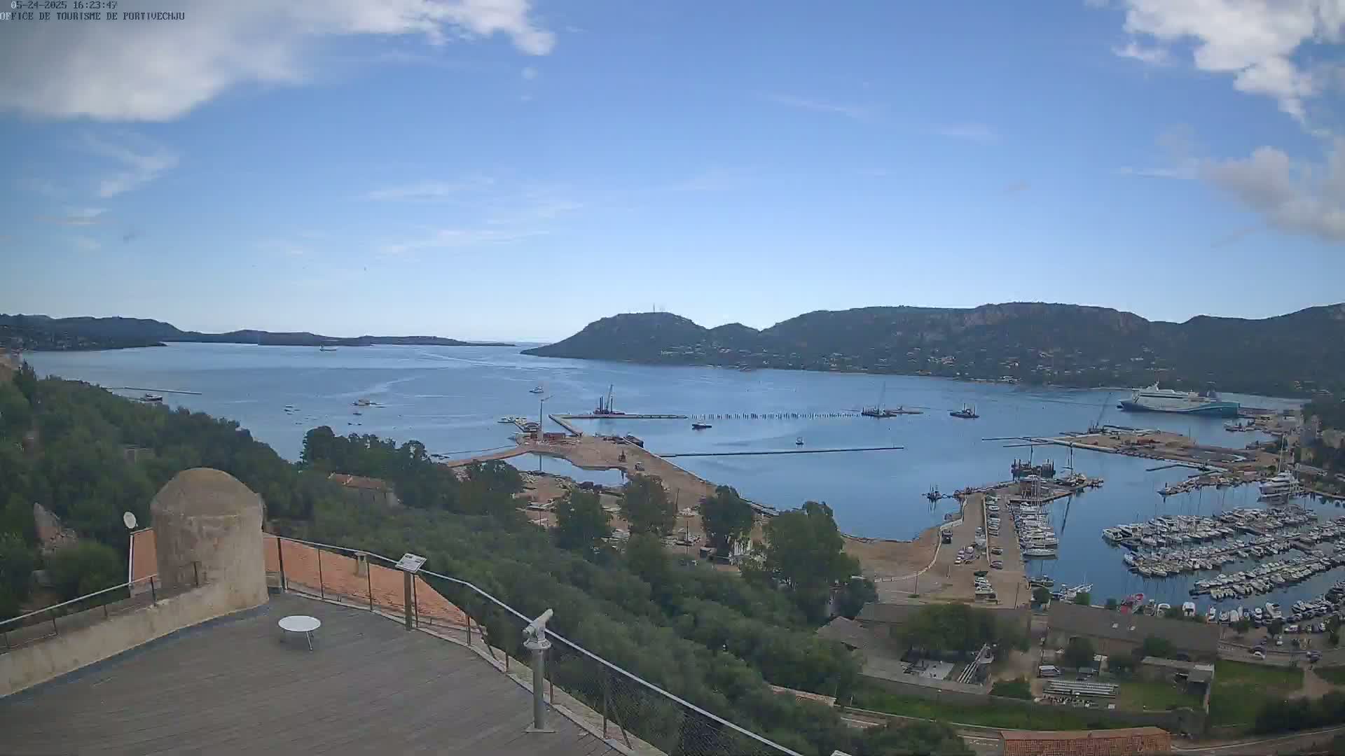 A partly cloudy day overlooks a harbor filled with boats and ships, with a larger body of water and hills in the distance.