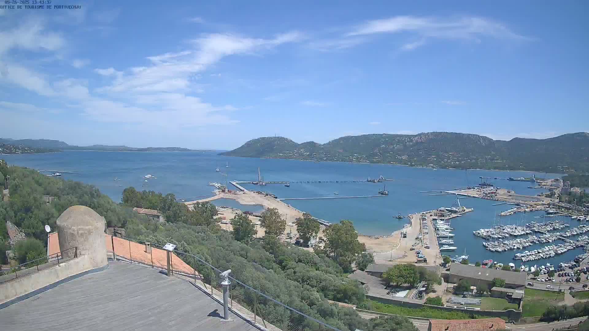 An elevated view of a harbor filled with boats, under a mostly sunny sky, with hills and a body of water in the background.