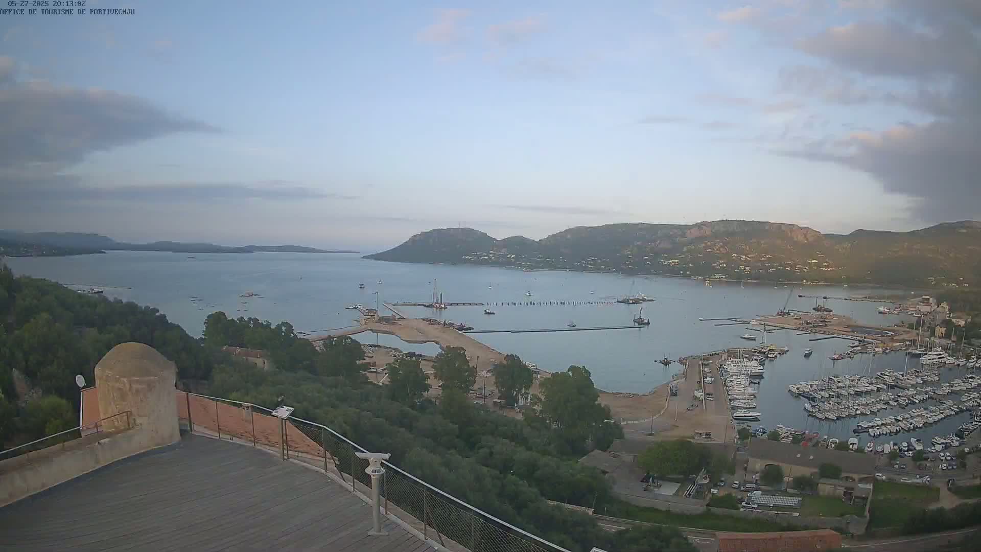 An expansive view of a harbor filled with numerous boats, nestled between hills under a mostly clear sky.
