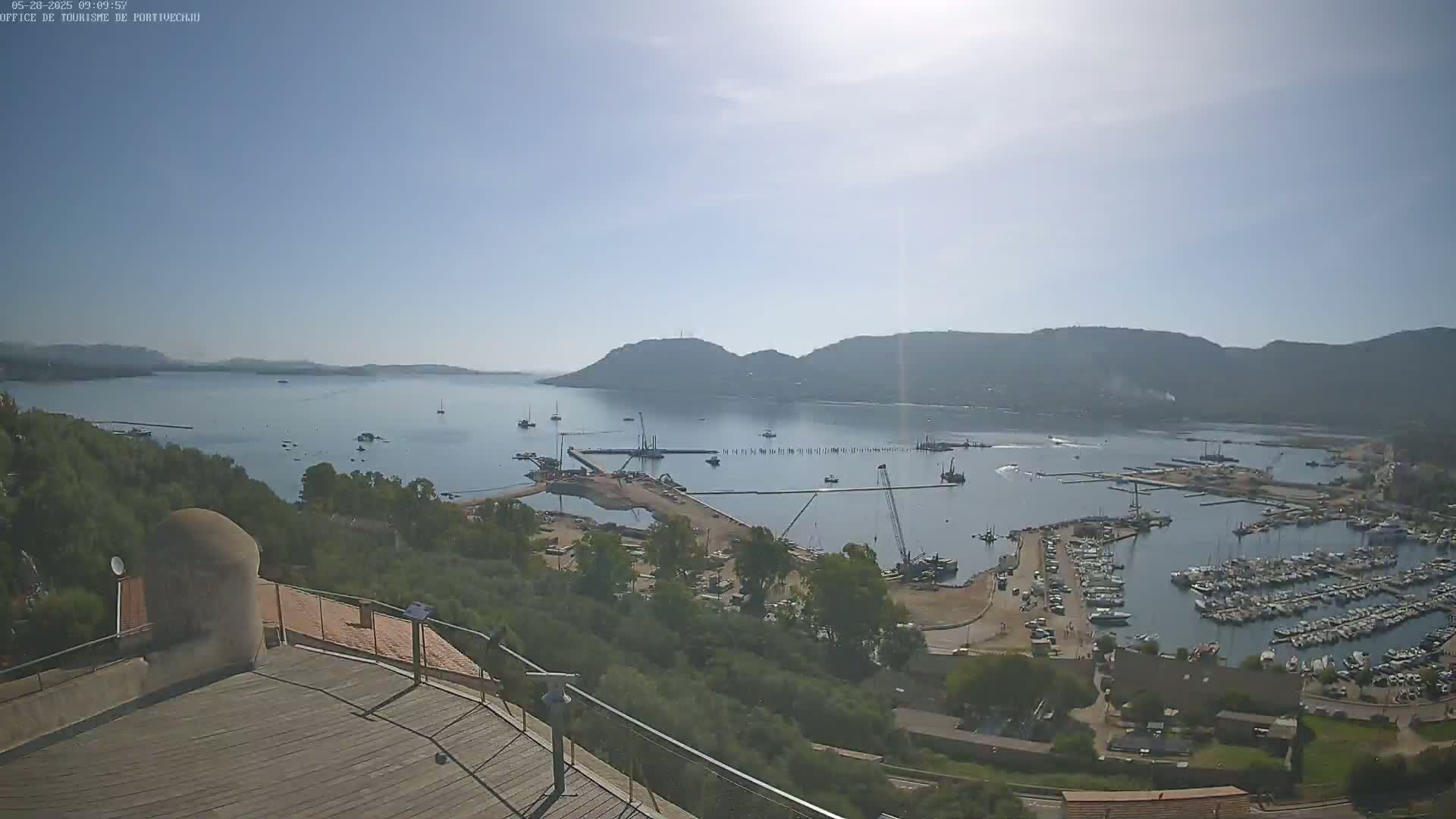 A sunny, high-angle view shows a harbor filled with boats, construction equipment, and surrounding hills, all overlooking a calm bay.