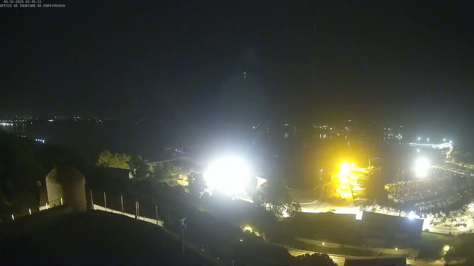 A nighttime aerial view shows brightly lit construction activity near a marina and body of water under a clear, dark sky.