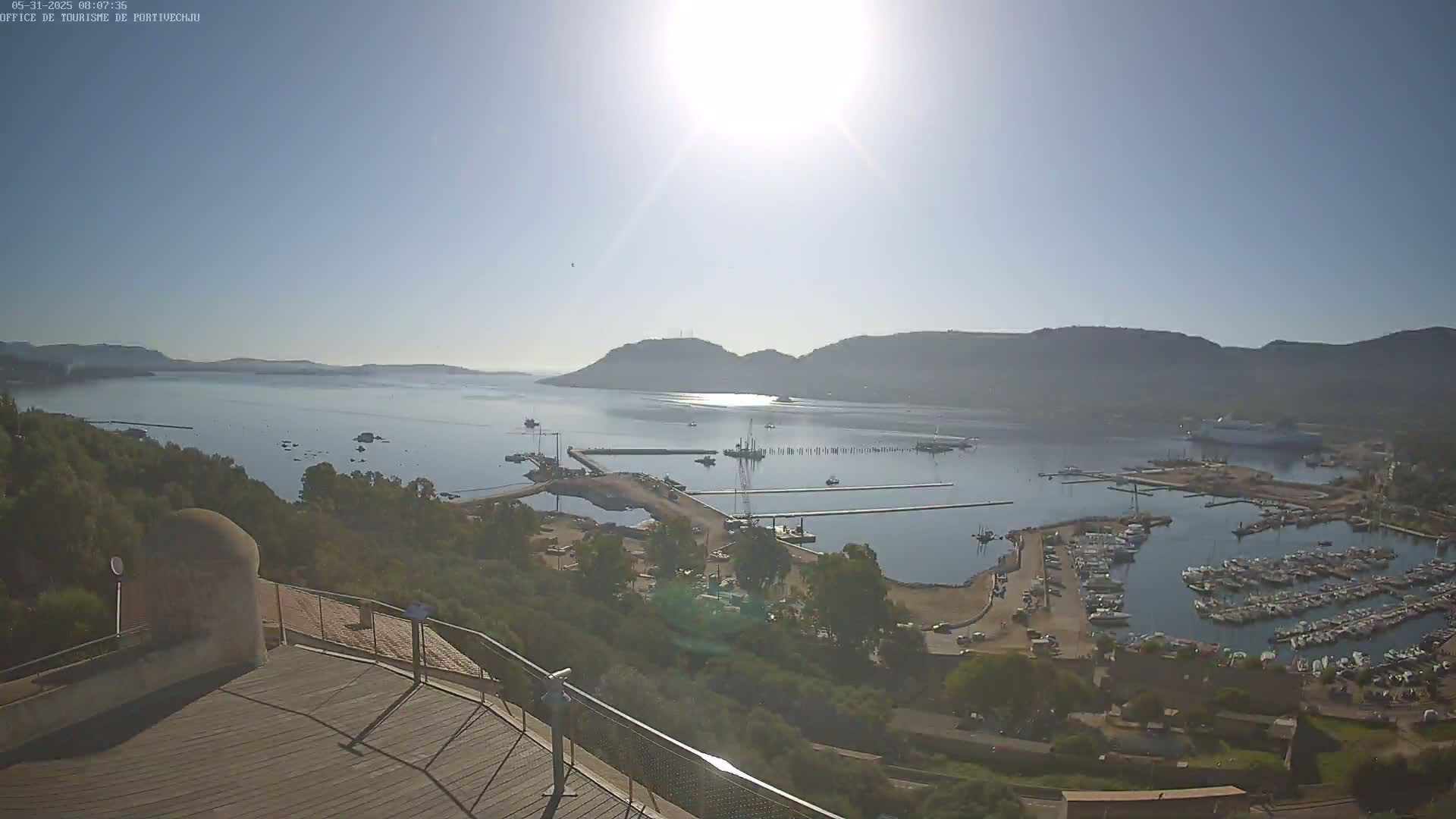 A sunny, hazy day overlooking a harbor filled with numerous boats and yachts, a large body of water, and hills in the distance.
