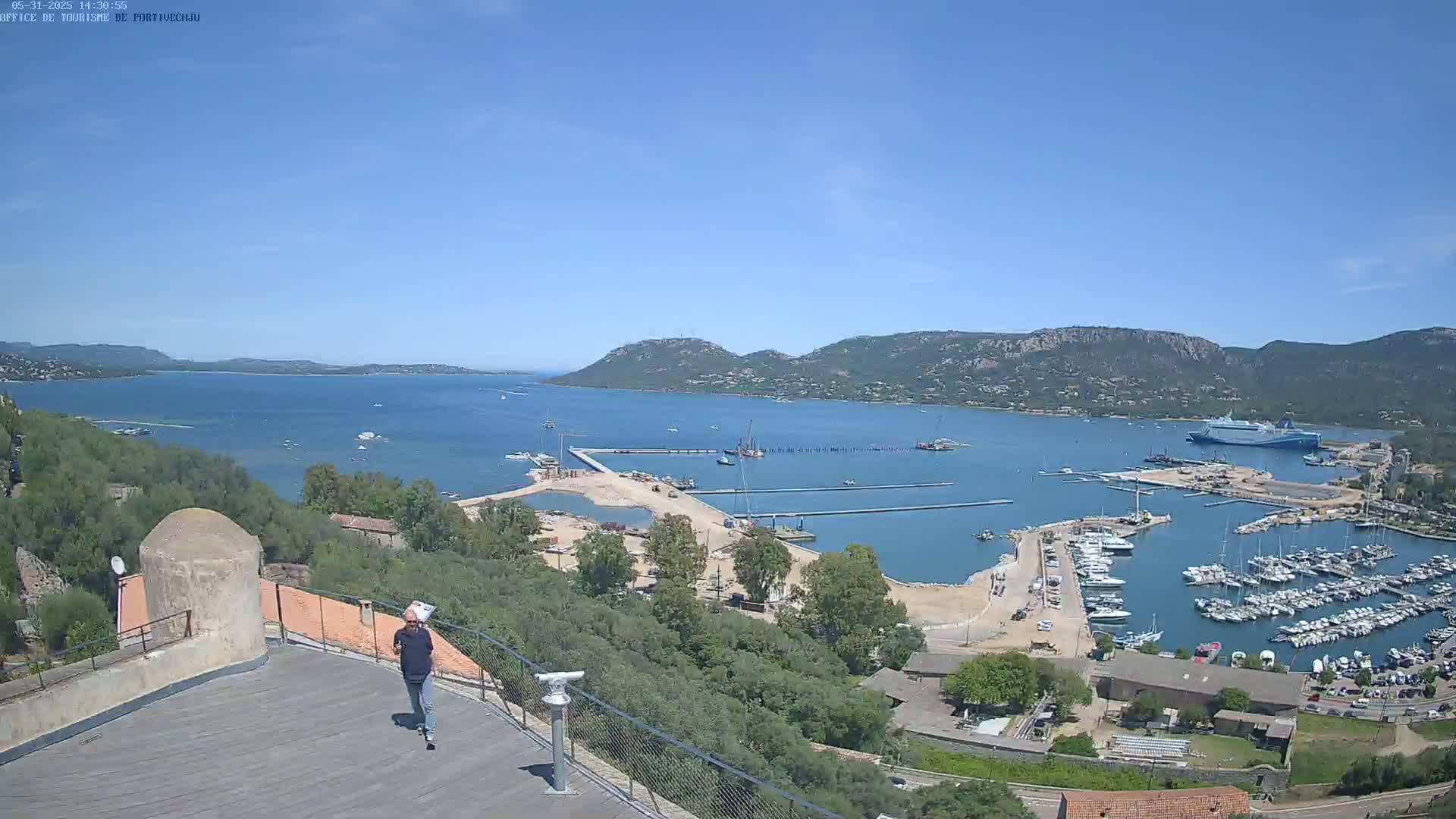 A man walks on a rooftop overlooking a harbor filled with boats and a large body of water under a clear blue sky.