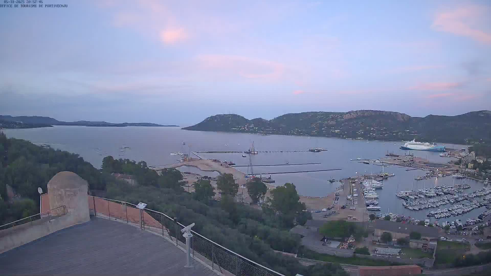 A high-angle, twilight view of a harbor filled with numerous boats and ships, nestled between land and a calm bay under a mostly clear sky.
