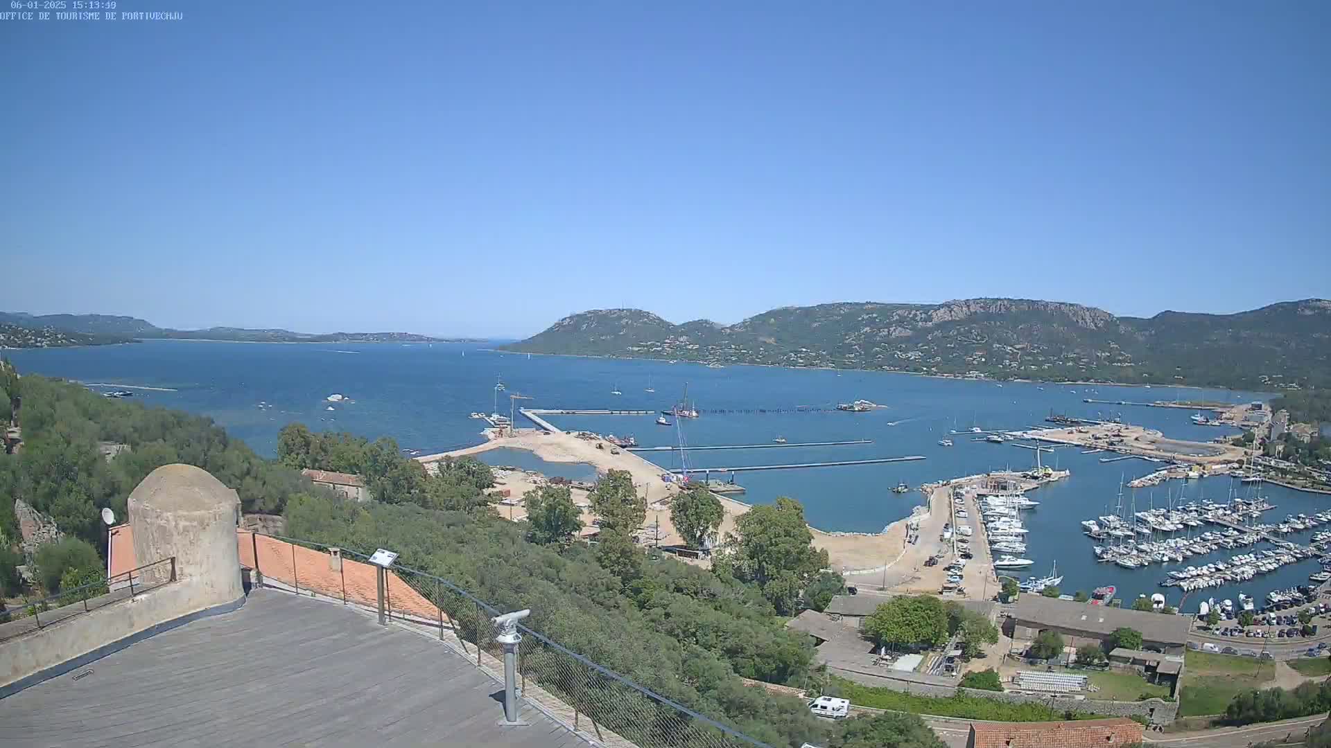 A high-angle, long shot on a clear, sunny day shows a marina filled with boats, construction along the shoreline, and a calm bay surrounded by hills.