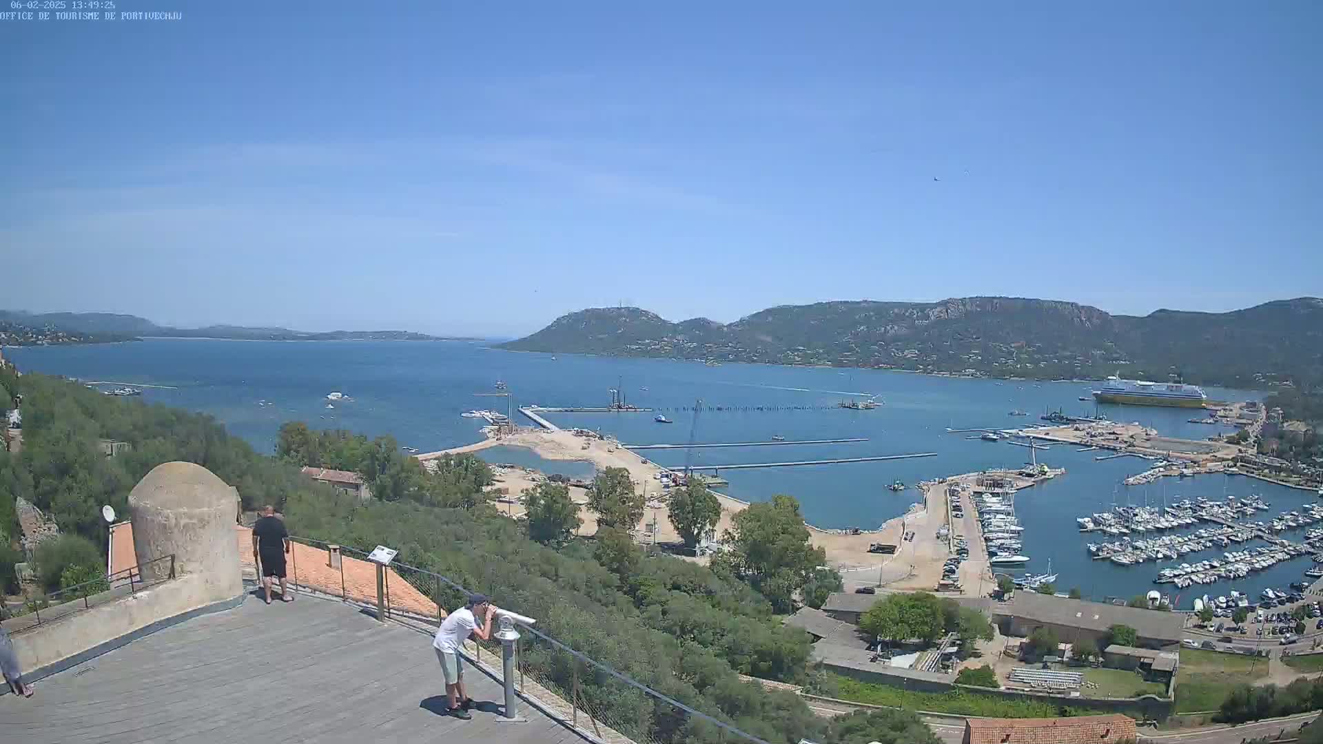 A sunny view overlooking a harbor filled with boats, a partially constructed pier, and a large body of water surrounded by hills.