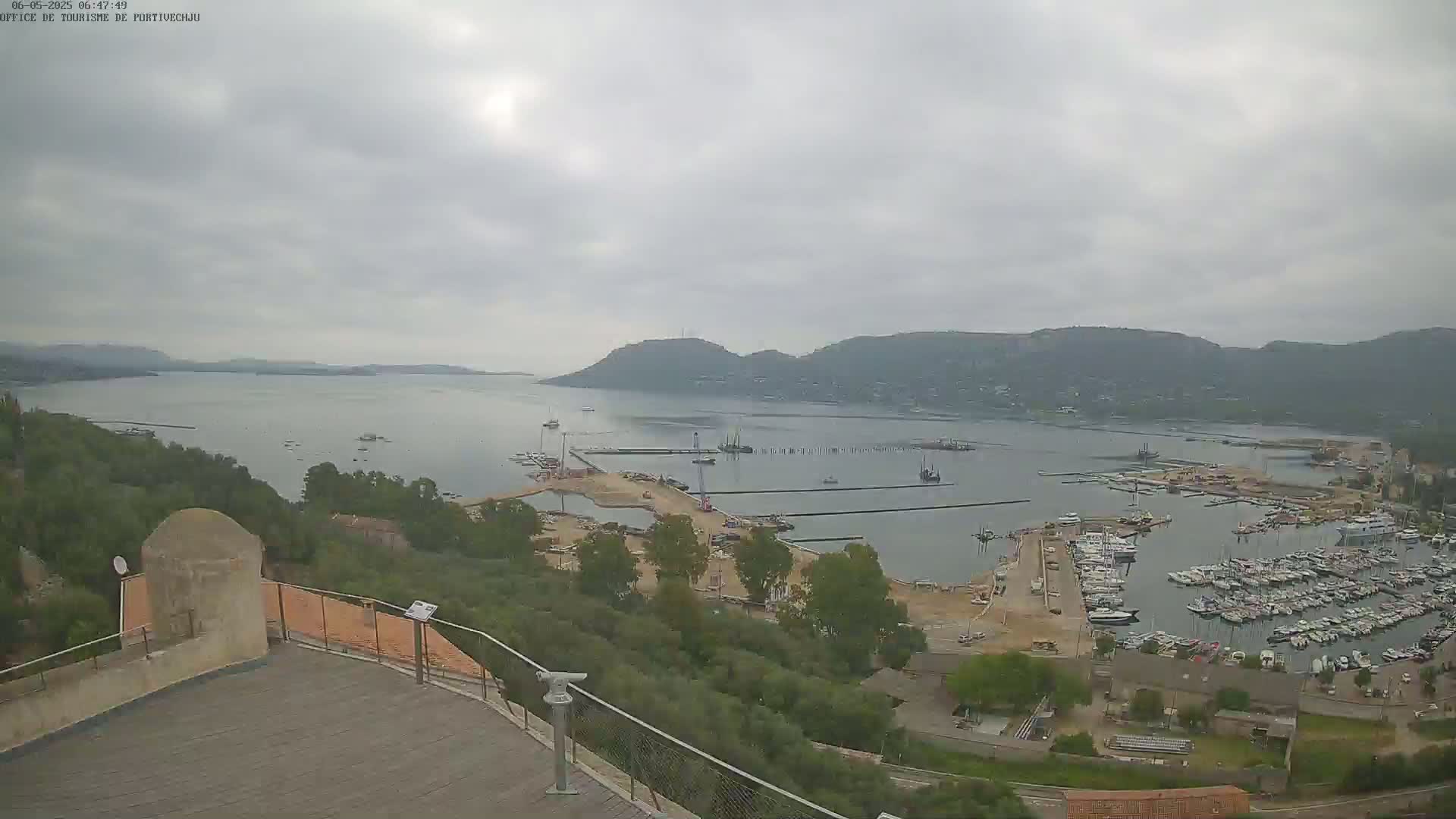 An overcast day reveals a harbor filled with boats, under construction, and surrounded by green hills and a calm body of water.