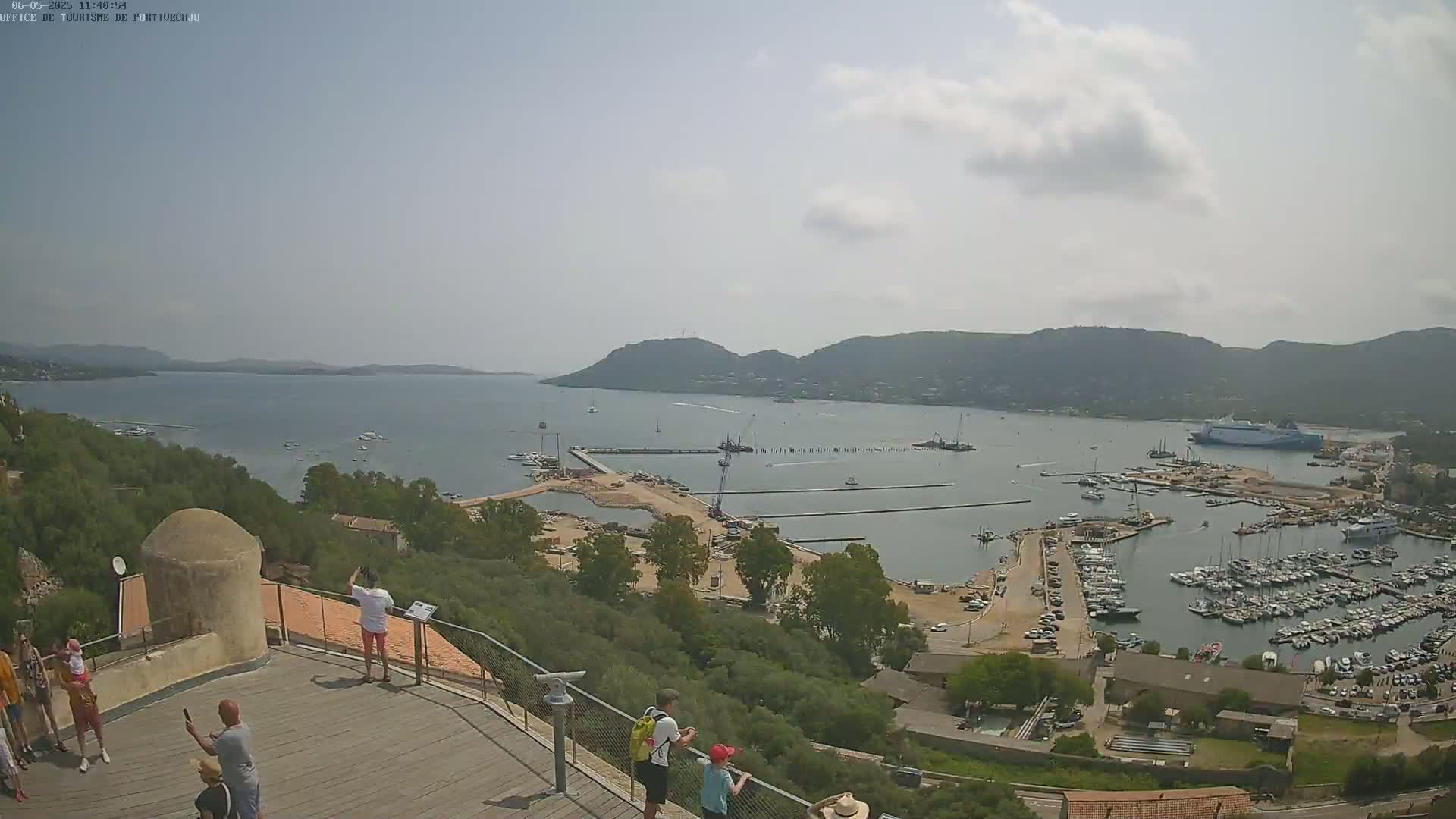 A high-angle, sunny view of a harbor filled with boats, construction, and surrounding hills, with several people standing on an elevated wooden platform.