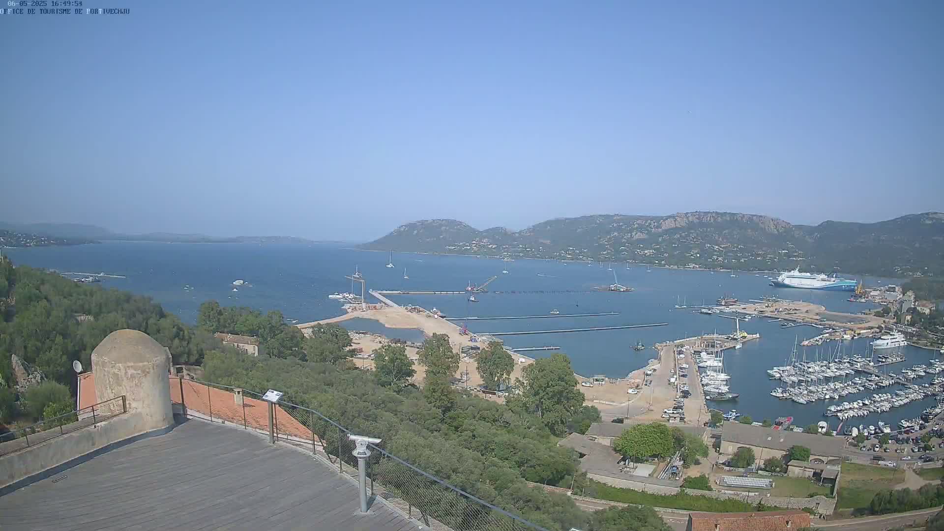 An aerial view on a clear, sunny day shows a harbor filled with numerous boats and yachts, under construction, nestled between green hills and a calm bay.