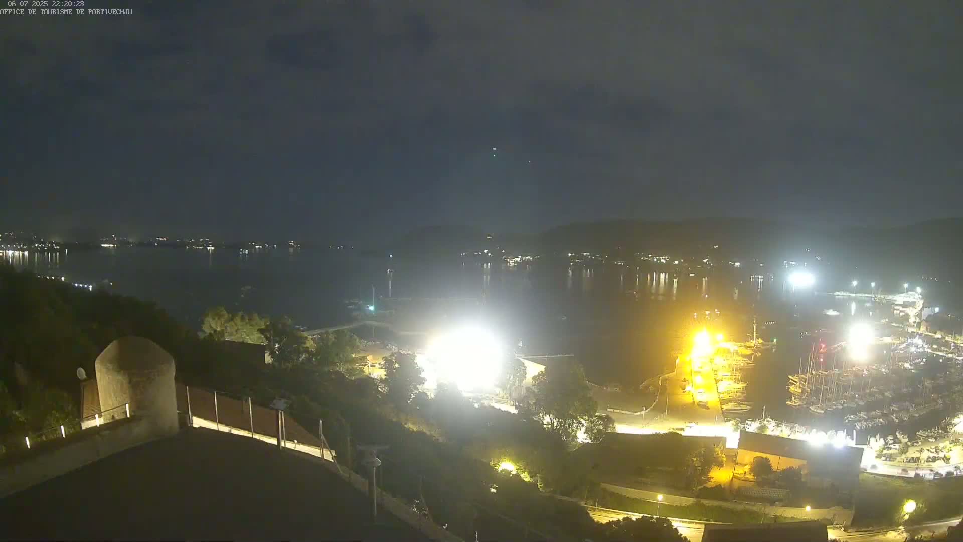 A nighttime view of a harbor filled with boats, brightly lit from the shore, overlooks a calm body of water under a dark, slightly cloudy sky.