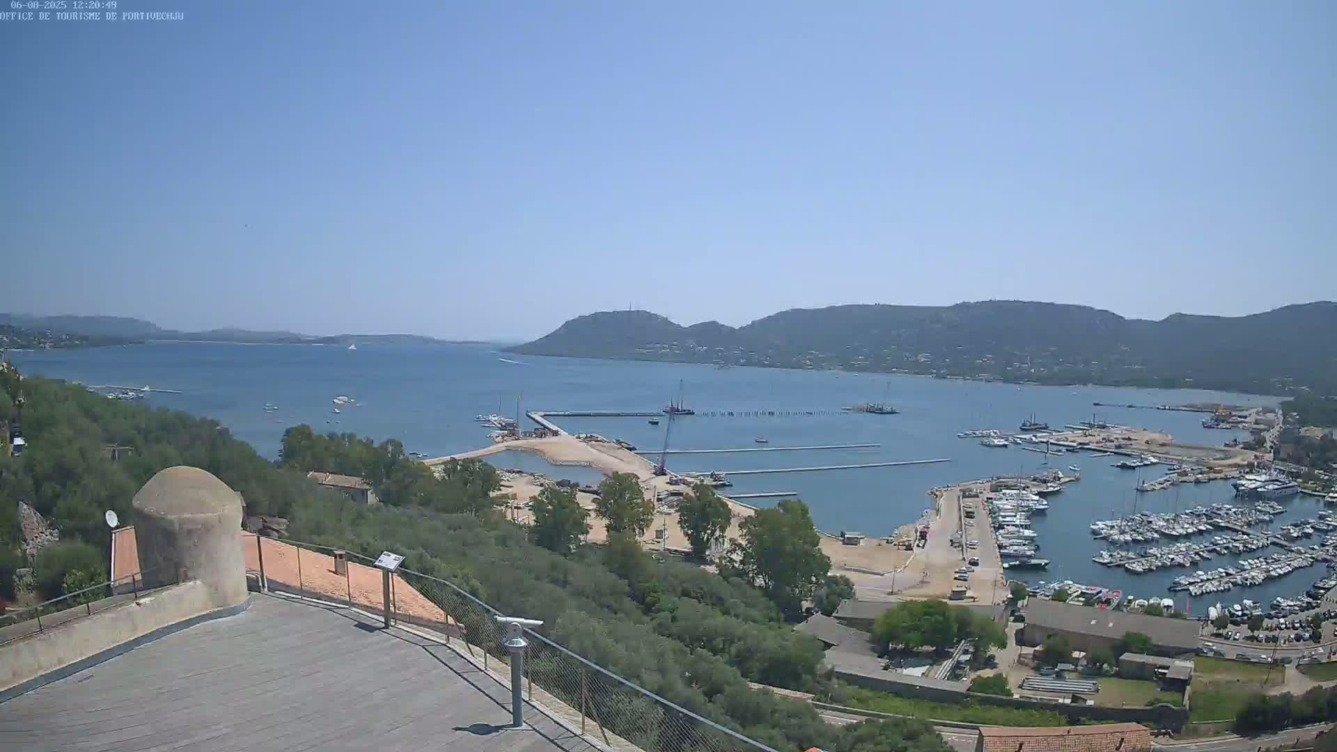 An elevated view of a harbor filled with numerous boats, under a clear blue sky, shows ongoing construction near the marina, and a hilly landscape in the background.
