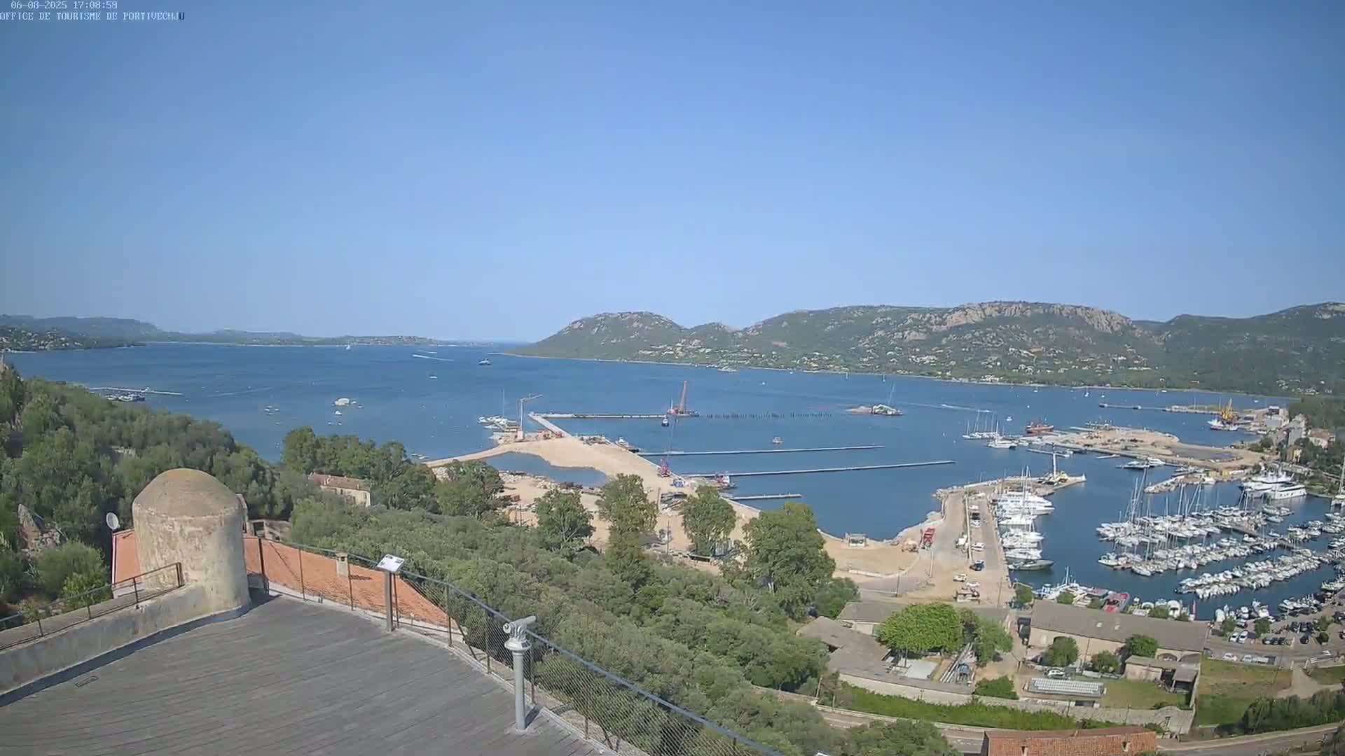 A harbor filled with boats is visible from an elevated vantage point on a clear, sunny day, with a bay and distant mountains in the background.