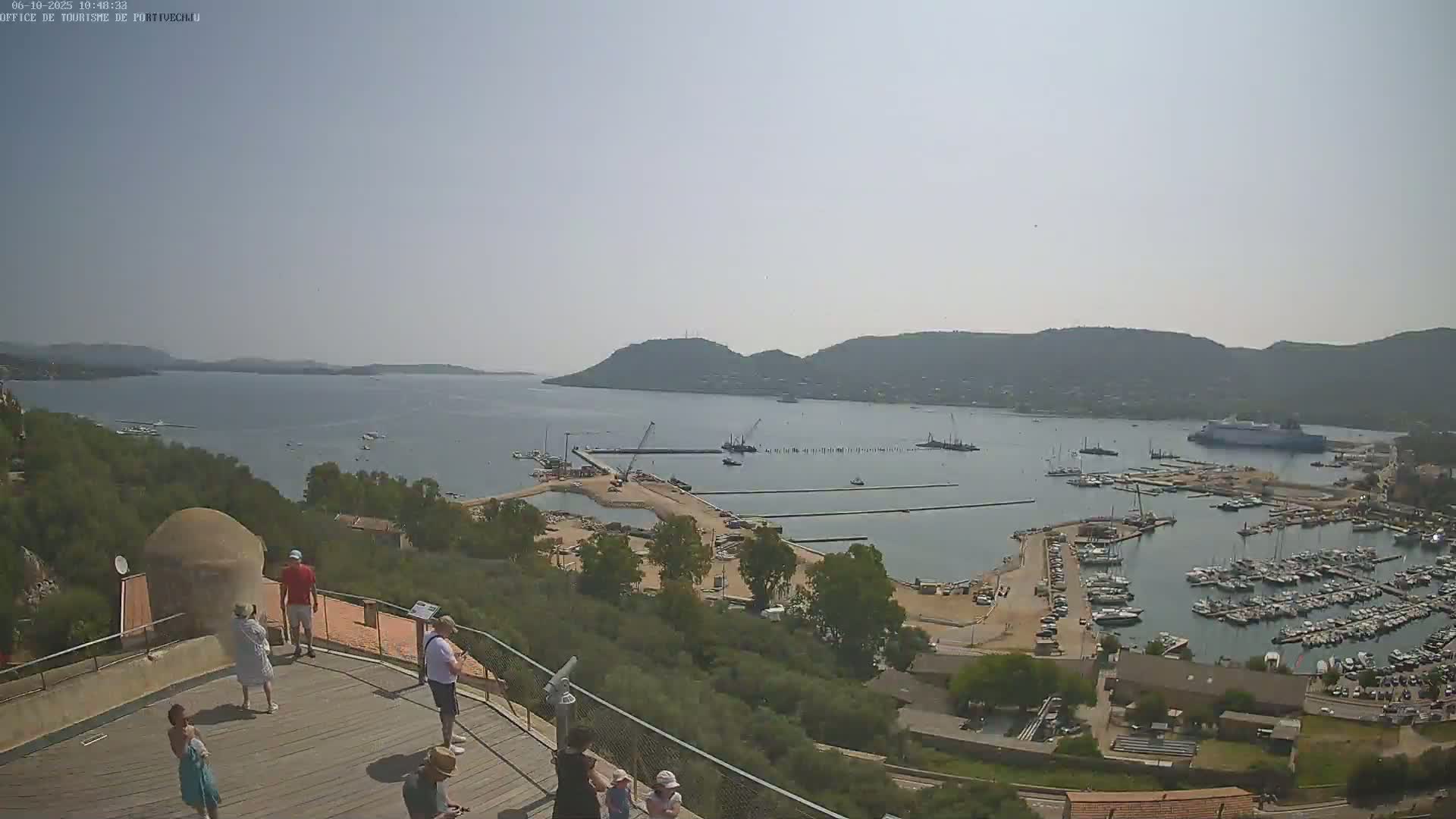An aerial view shows a harbor filled with boats on a sunny day, with tourists observing the scene from an elevated vantage point.