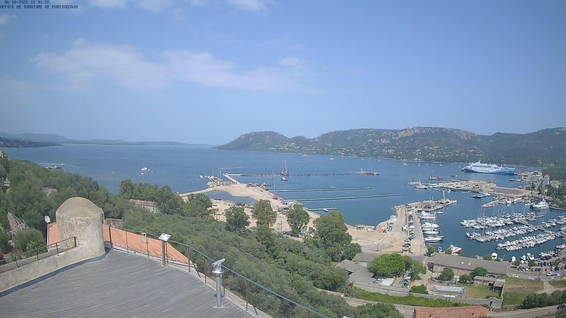 An elevated view of a harbor filled with boats, under a mostly clear sky, shows a large body of water extending to distant hills.