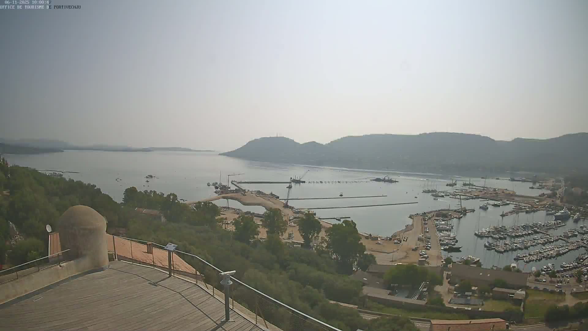 A hazy, high-angle view shows a marina filled with boats and construction activity near a calm bay surrounded by hills.