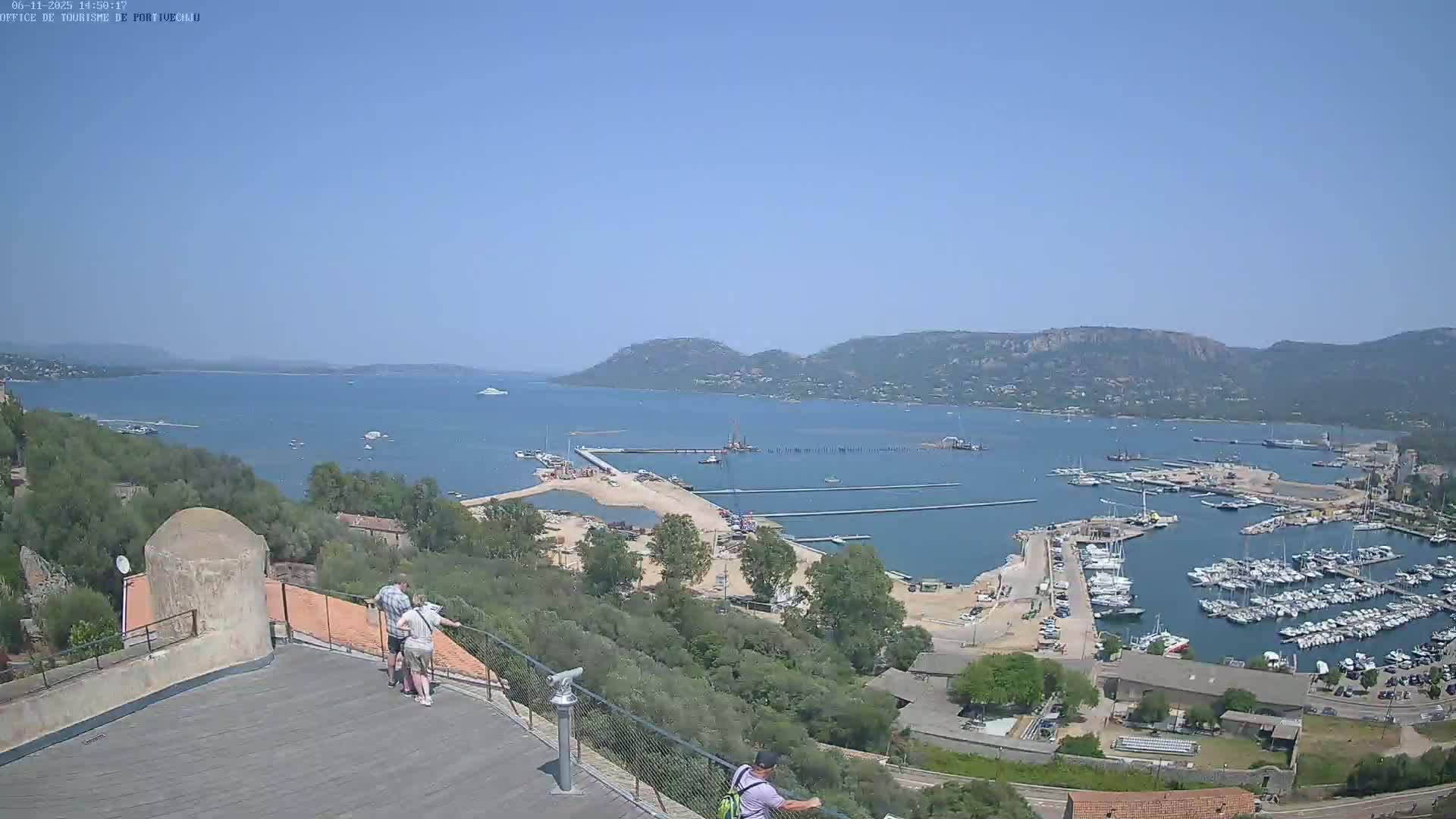A high-angle, sunny view shows a harbor filled with boats, construction activity, and a calm bay under a clear blue sky.